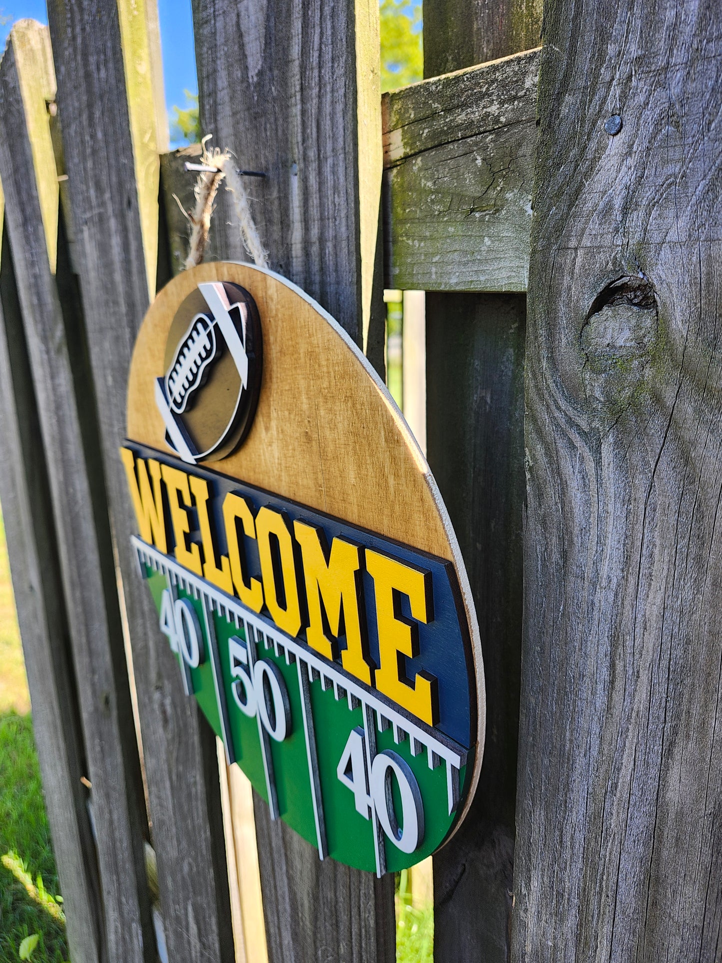 Round wood door sign hanging from a fence with twine. The sign's background has a light colored stain. At the top of the sign is a brown football with white laces. Below that is "Welcome" in bold yellow letters with a navy blue background. Below that (at the bottom of the sign) is a green semi-circle with white yard lines marking "40", "50", "40". The sign is at a slight angle at this photo showing the 3D dimension.