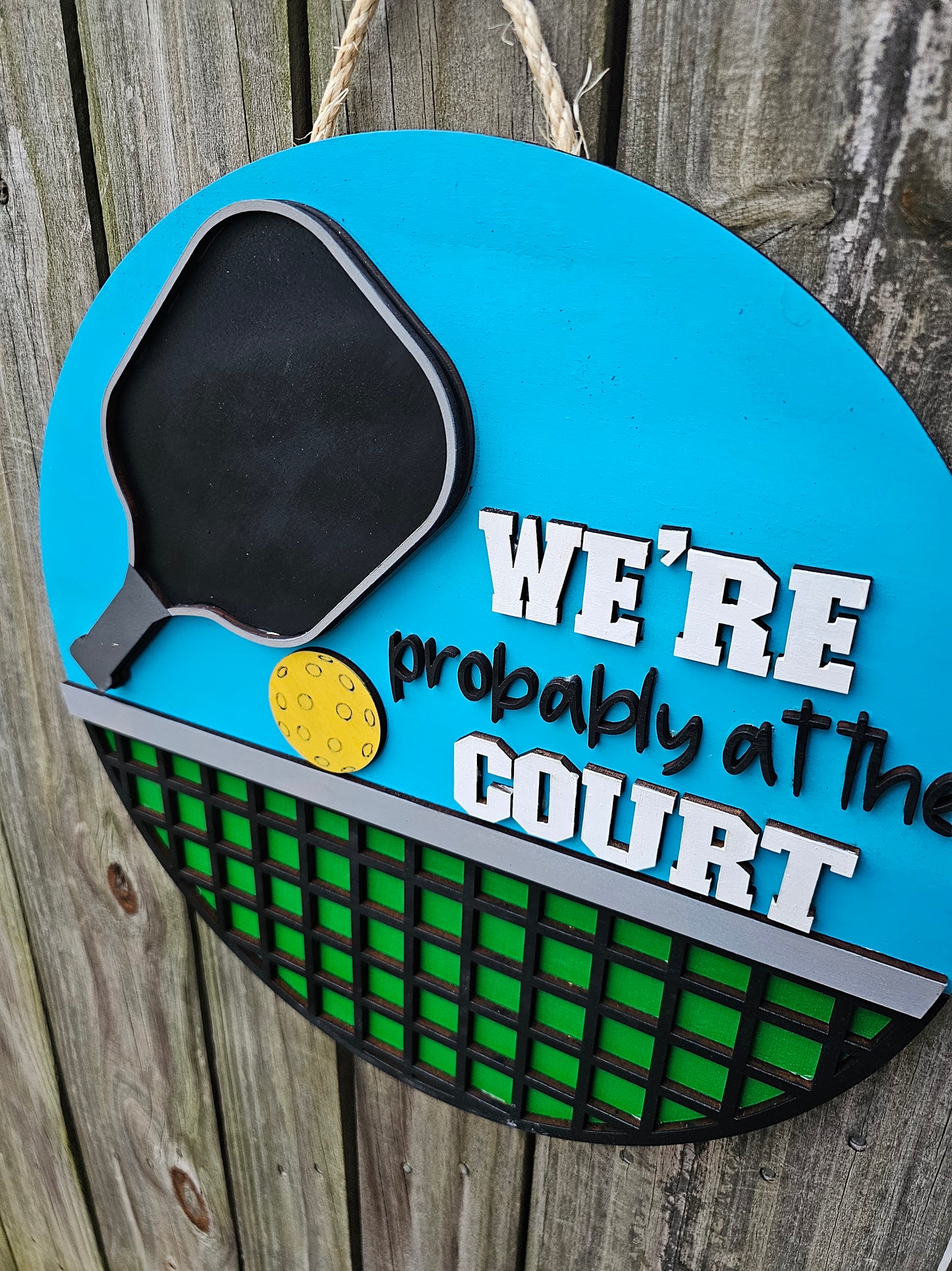 Round wood pickleball themed door sign hanging on fence. The sign has a bright blue background on top with a pickleball paddle, ball, and "We're Probably At the Court." The bottom portion of the sign is green with black net. The sign is slightly angled in this photo showing 3D elements.