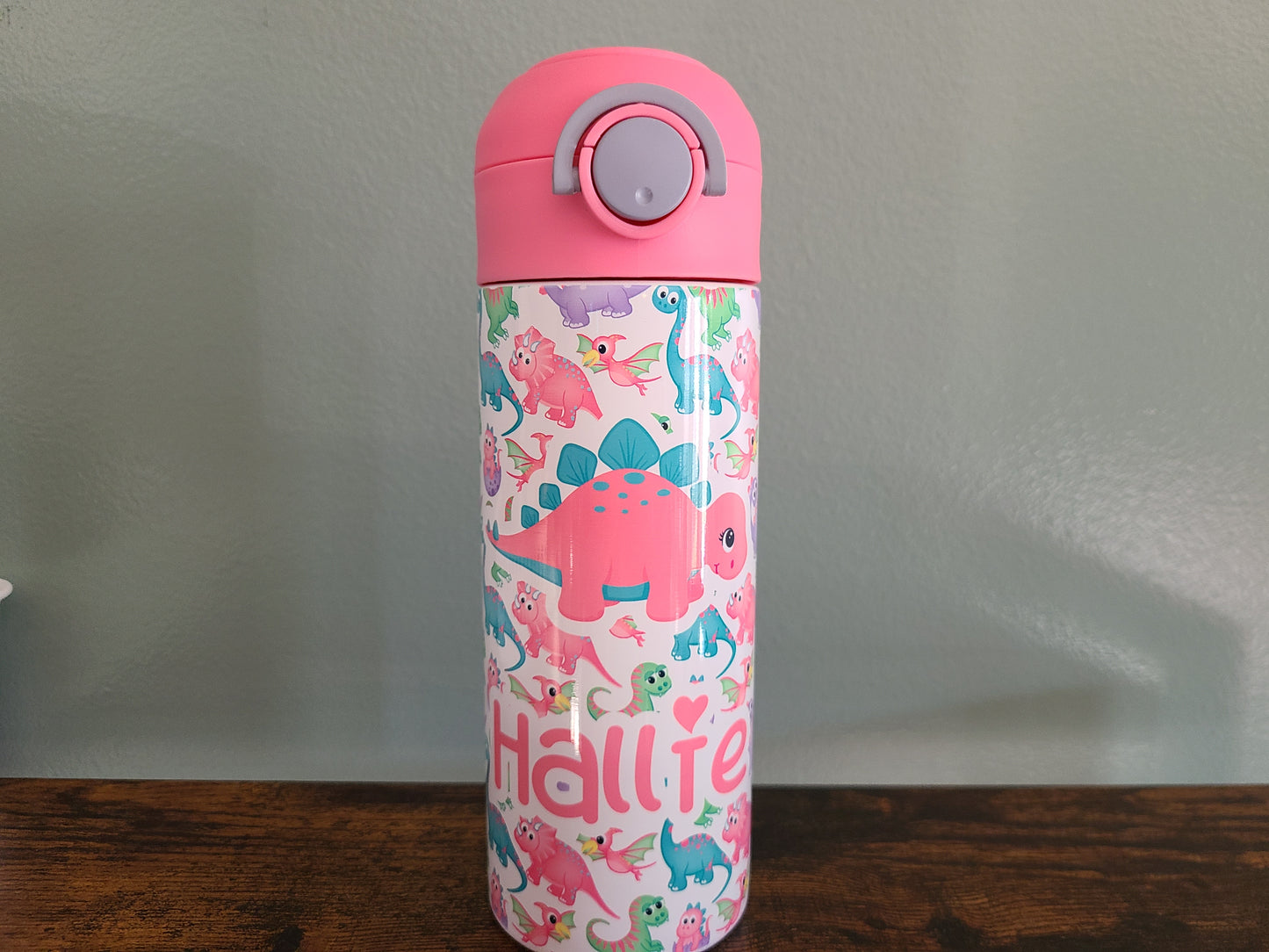 12 oz water bottle with pink lid that has a built in straw, locking push button and handle. This design features pastel rainbow dinosaurs. Personalized in coordinating colors. Back to School.