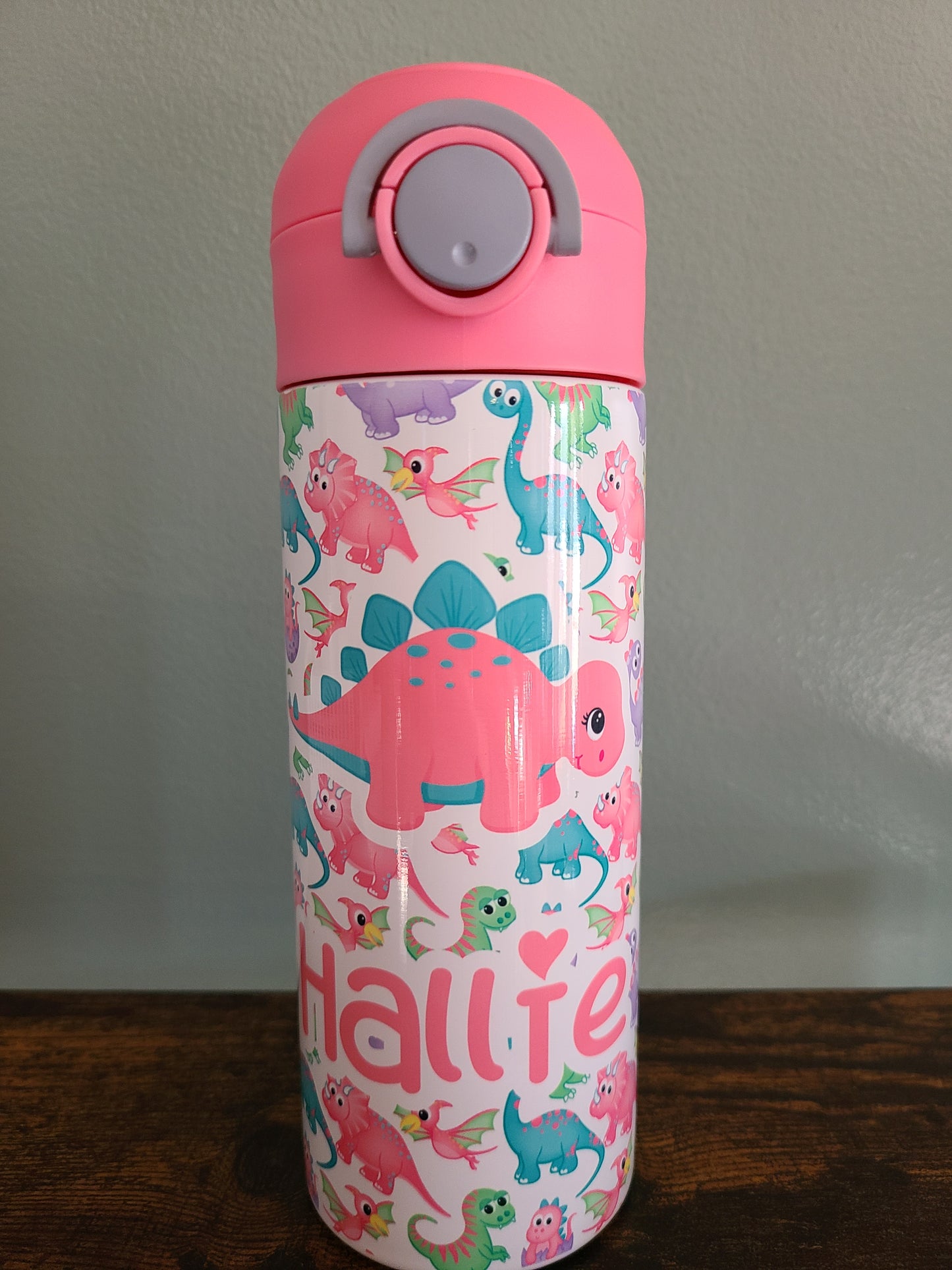 12 oz water bottle with pink lid that has a built in straw, locking push button and handle. This design features pastel rainbow dinosaurs. Personalized in coordinating colors. Back to School.
