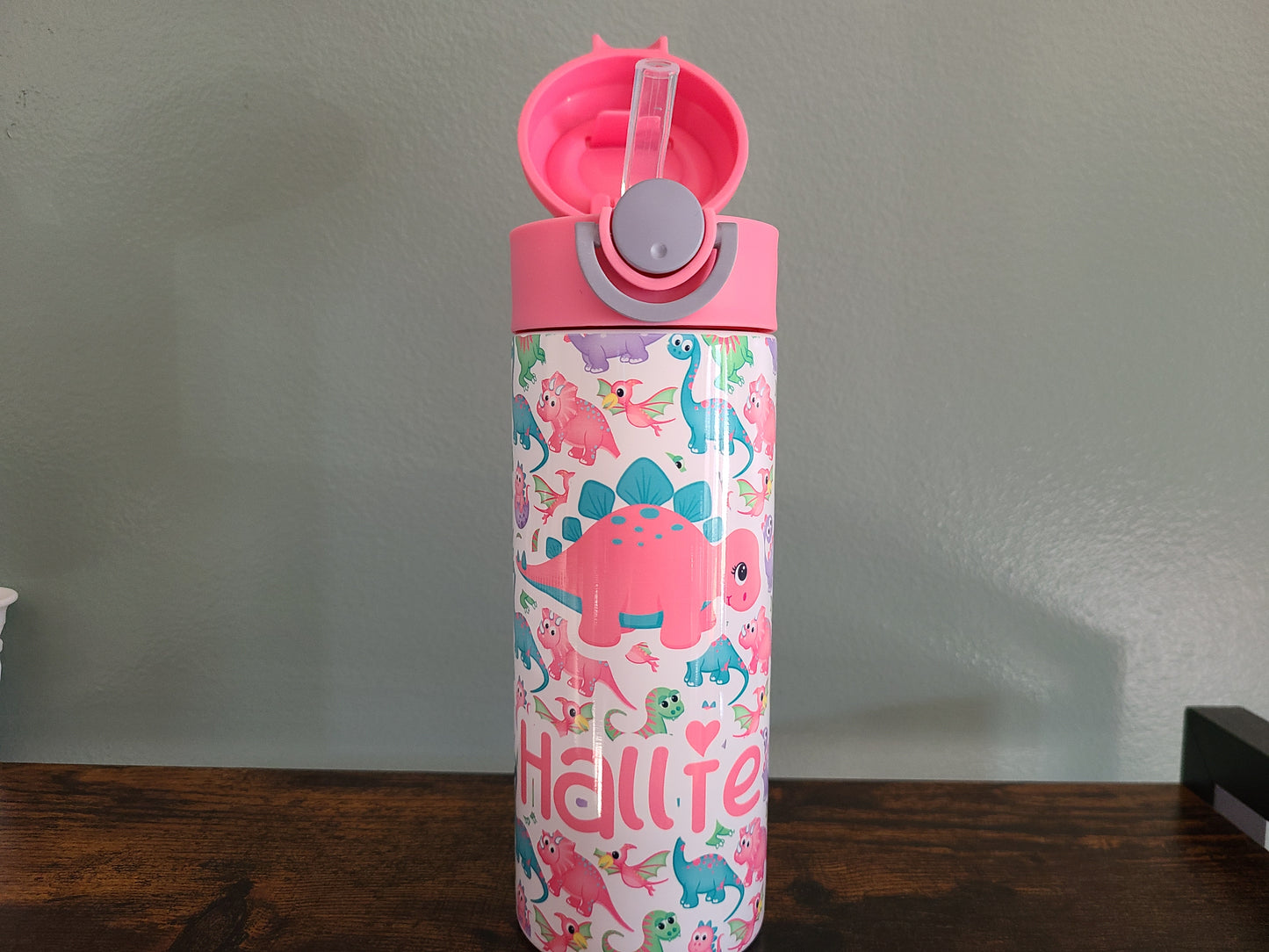 12 oz water bottle with pink lid that has a built in straw, locking push button and handle. This design features pastel rainbow dinosaurs. Personalized in coordinating colors. Back to School. Lid is open showing silicon straw