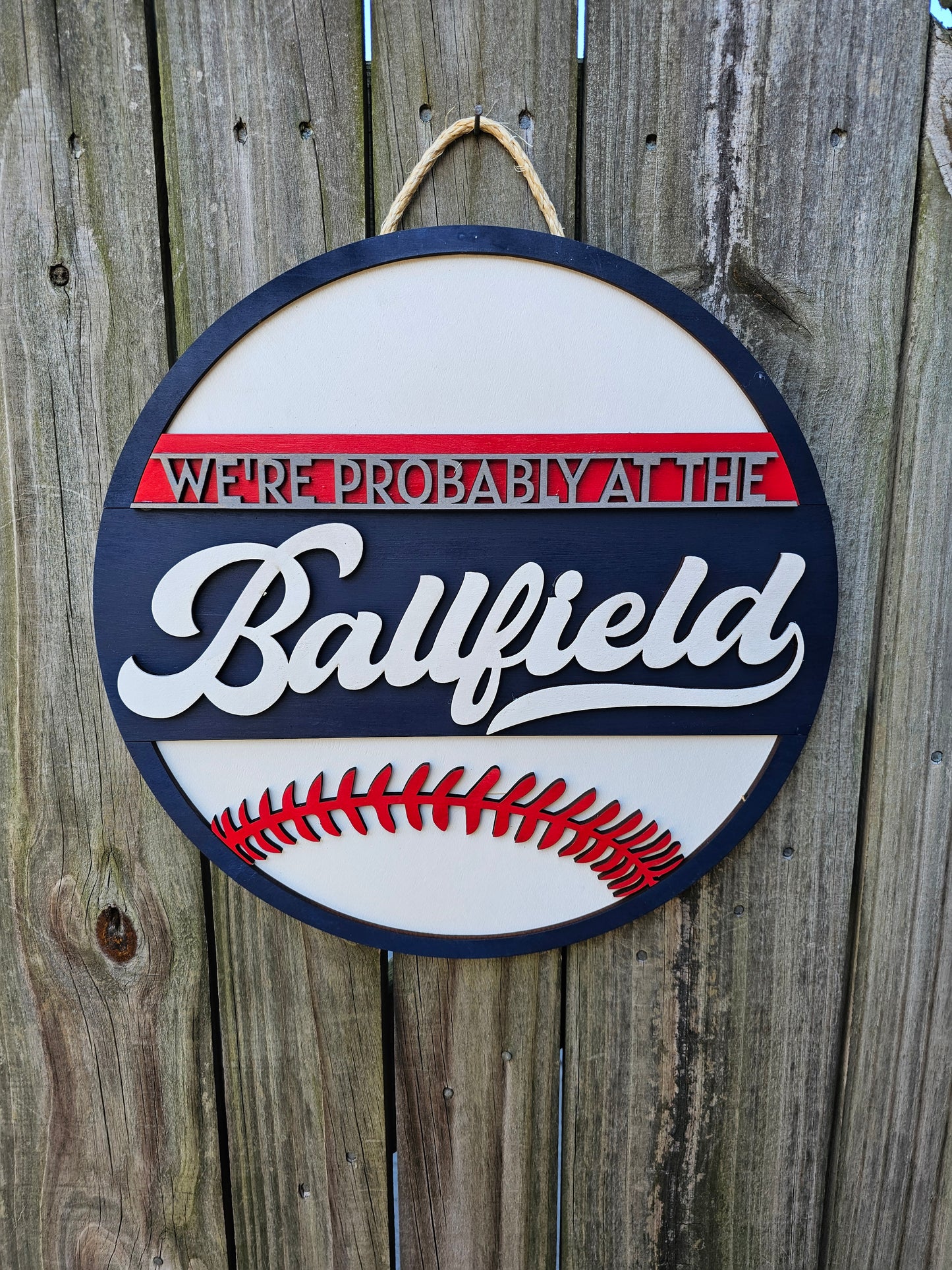 Round wood sign hanging on fence with "We're Probably At the Ballfield" and baseball laces. Sign is painted navy blue, red, and white. In this photo the sign is forward facing.