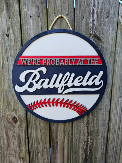 Round wood sign hanging on fence with "We're Probably At the Ballfield" and baseball laces. Sign is painted navy blue, red, and white. In this photo the sign is forward facing.