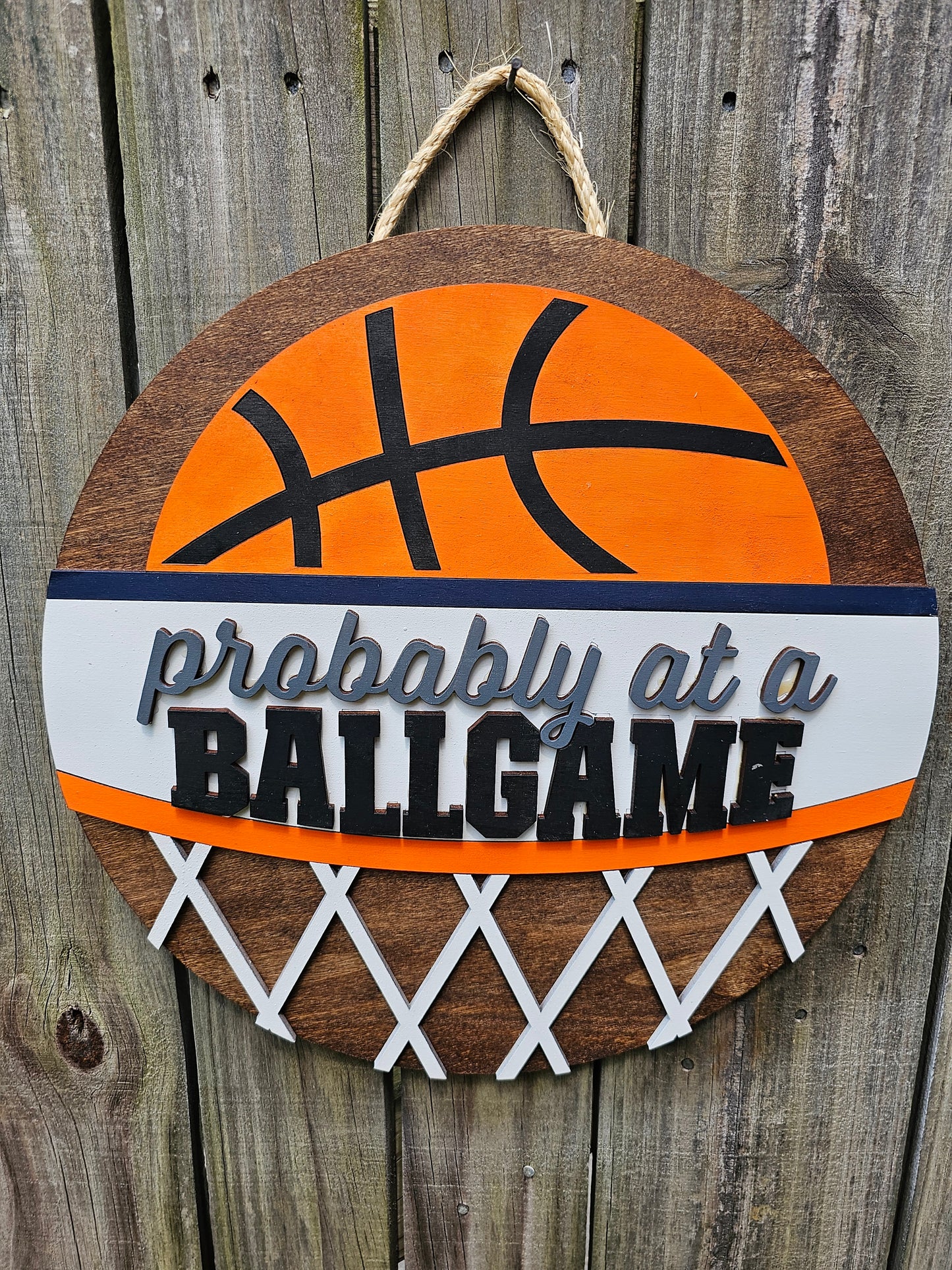 Round wood door sign with basketball design and text 'Probably at a BALLGAME'. Sign is hanging by twine on a wood fence. 