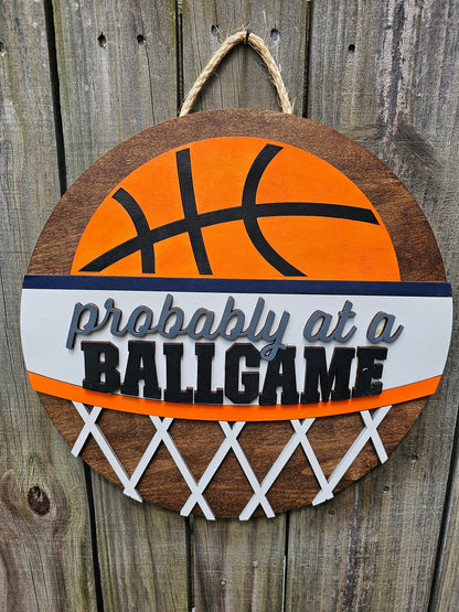 Round wood door sign with basketball design and text 'Probably at a BALLGAME'. Sign is hanging by twine on a wood fence. 
