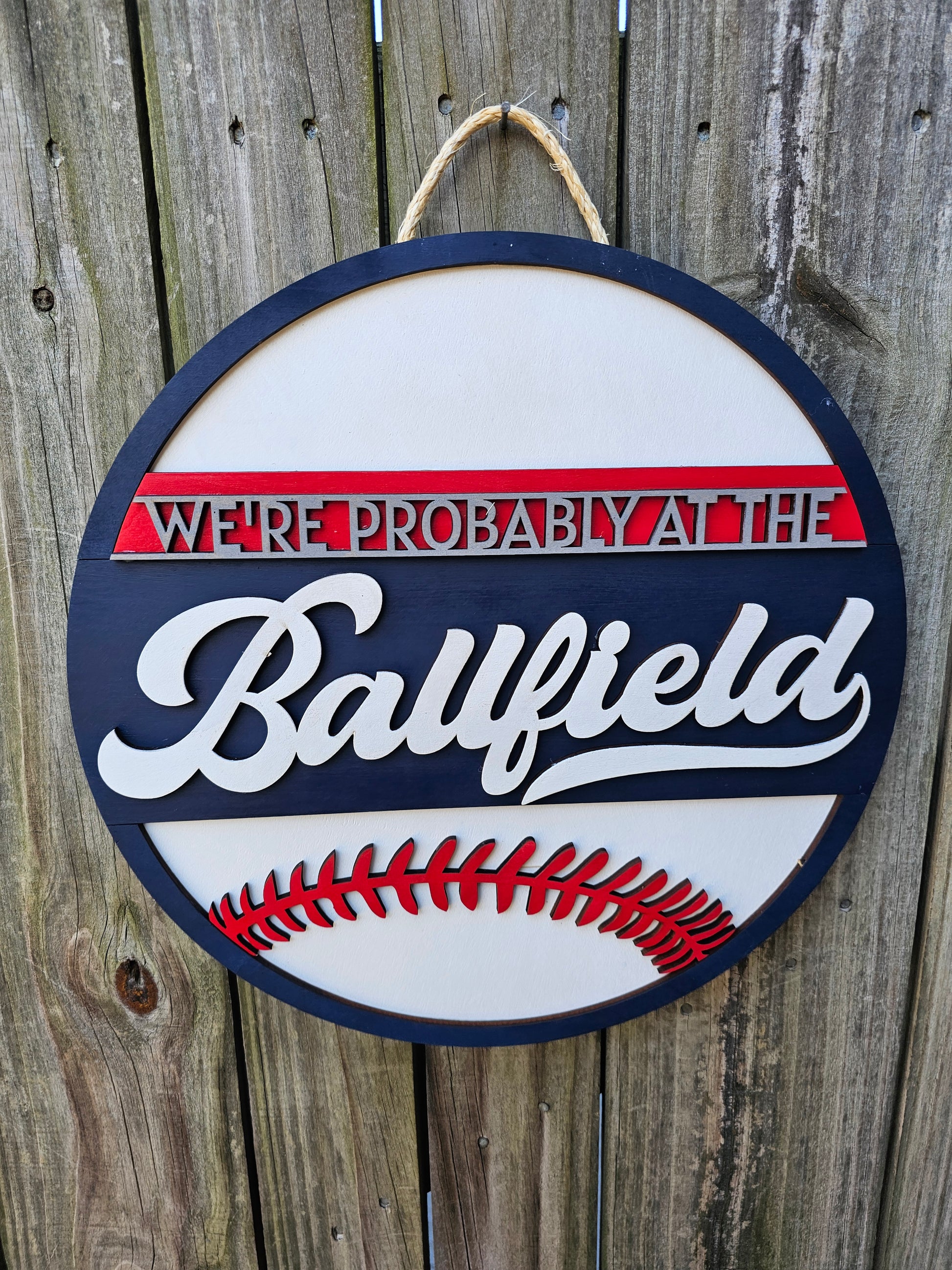 Round wood sign hanging on fence with "We're Probably At the Ballfield" and baseball laces. Sign is painted navy blue, red, and white. In this photo the sign is forward facing.