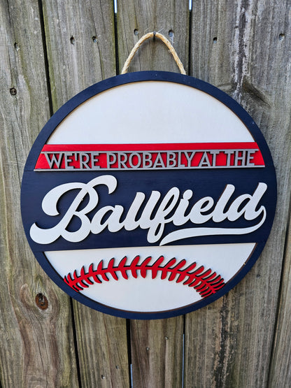 Round wood sign hanging on fence with "We're Probably At the Ballfield" and baseball laces. Sign is painted navy blue, red, and white. In this photo the sign is forward facing.