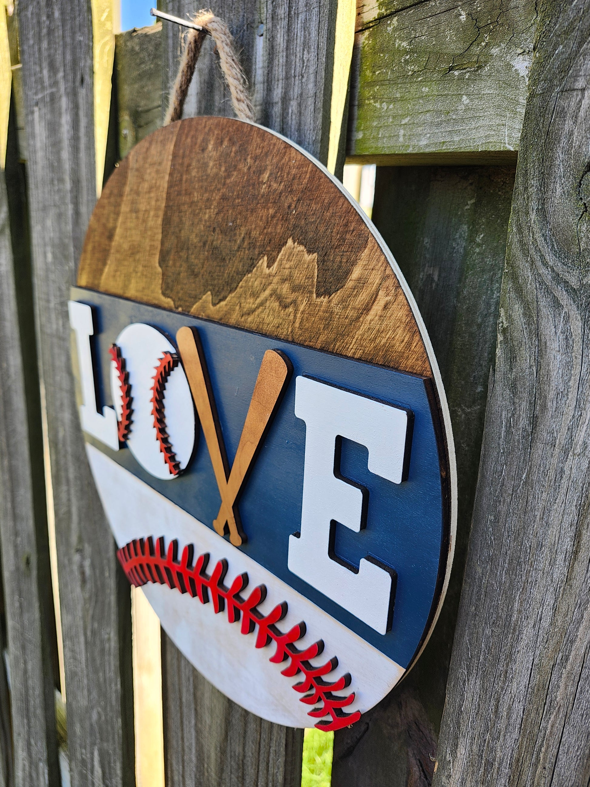 A round wooden door hanging sign with a baseball design, featuring the word 'LOVE' with the 'V' formed by two crossed baseball bats, painted in white, blue, and red colors. The photo is taken on a slight angle showing off the 3D details on the sign.