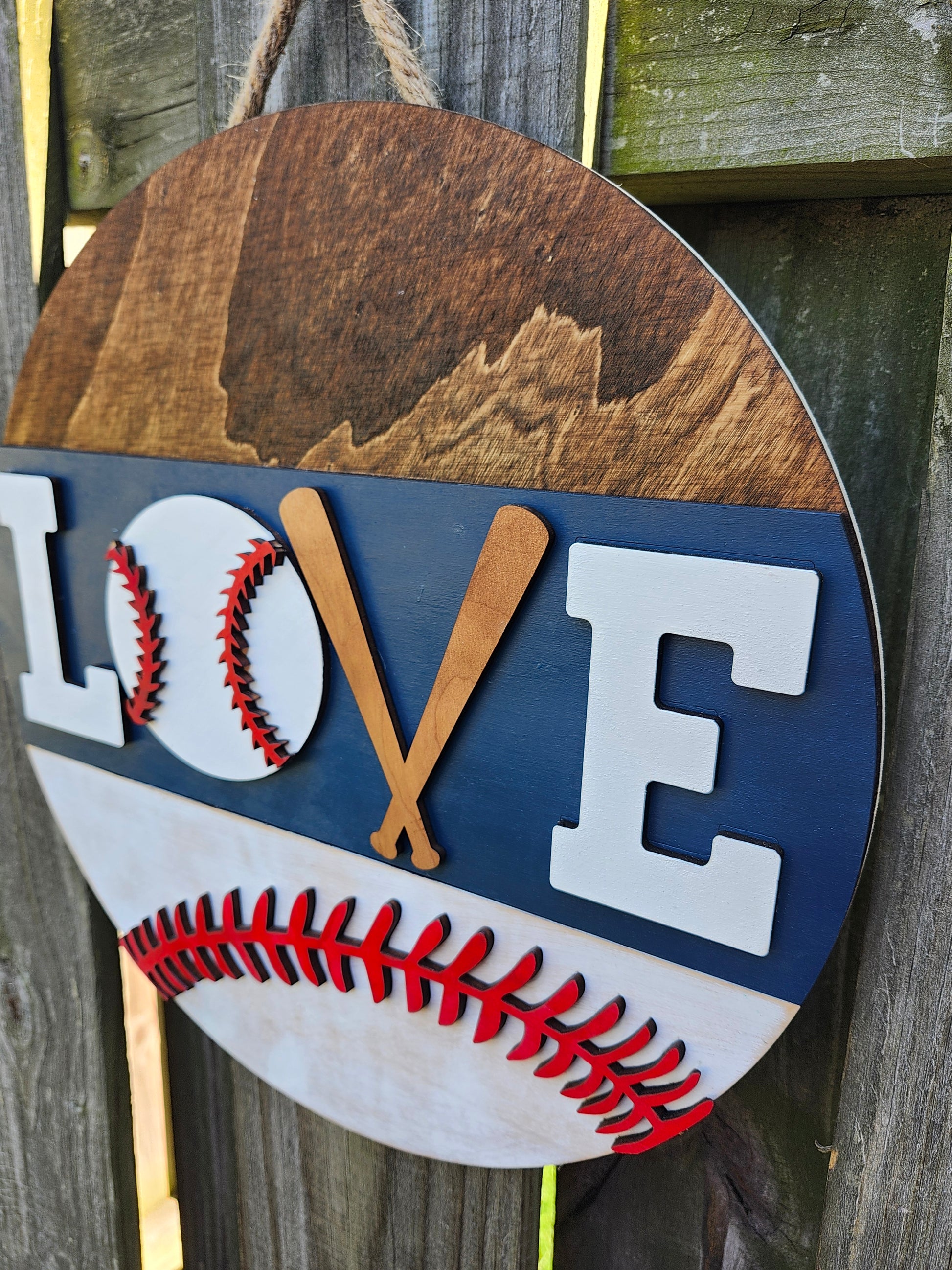 A round wooden door hanging sign with a baseball design, featuring the word 'LOVE' with the 'V' formed by two crossed baseball bats, painted in white, blue, and red colors. The photo is taken on a slight angle showing off the 3D details on the sign.