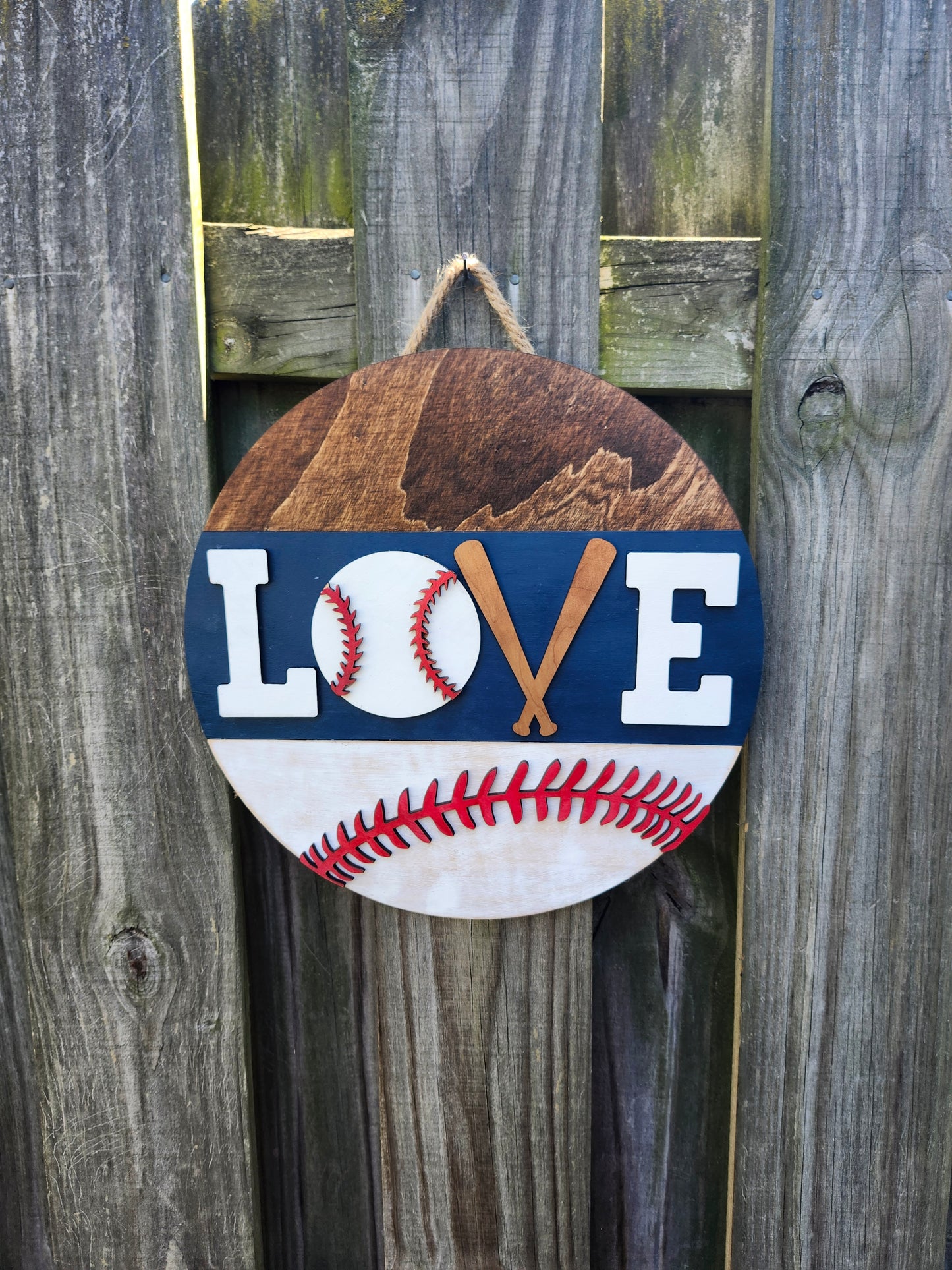 A round wooden door hanging sign with a baseball design and the word 'LOVE' where the 'V' is formed by two crossed baseball bats, hanging on a wooden fence. The sign is painted navy blue, white and red.