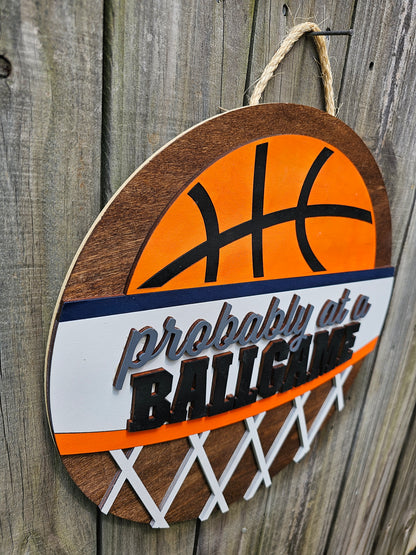 Round wood door sign with basketball design and text 'Probably at a BALLGAME'. Sign is hanging by twine on a wood fence. The photo is taken from an angle showing the 3D dimensions of the sign.