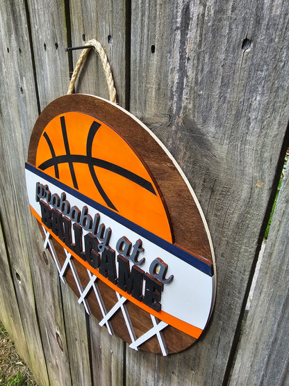 Round wood door sign with basketball design and text 'Probably at a BALLGAME'. Sign is hanging by twine on a wood fence. The photo is taken from an angle showing the 3D dimensions of the sign.