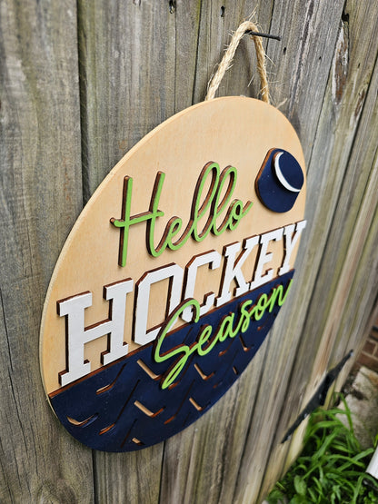 Round wooden sign with 'Hello Hockey Season' text and hockey puck design on a wooden background. This photo is taken at an angle showing off 3D elements.