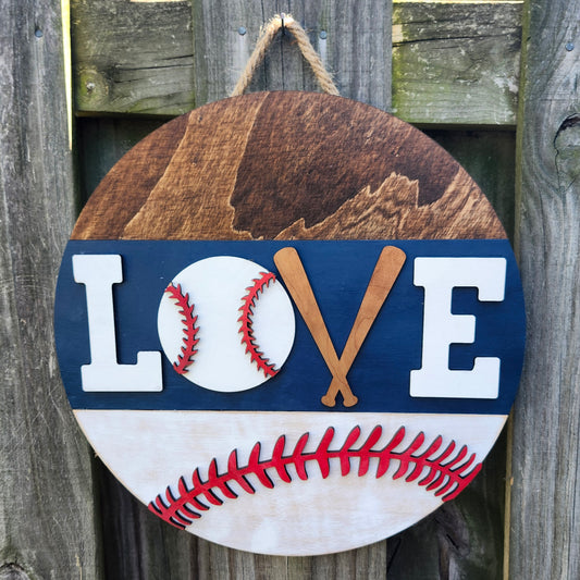 A round wooden door hanging sign with a baseball design, featuring the word 'LOVE' with the 'V' formed by two crossed baseball bats, painted in white, blue, and red colors.