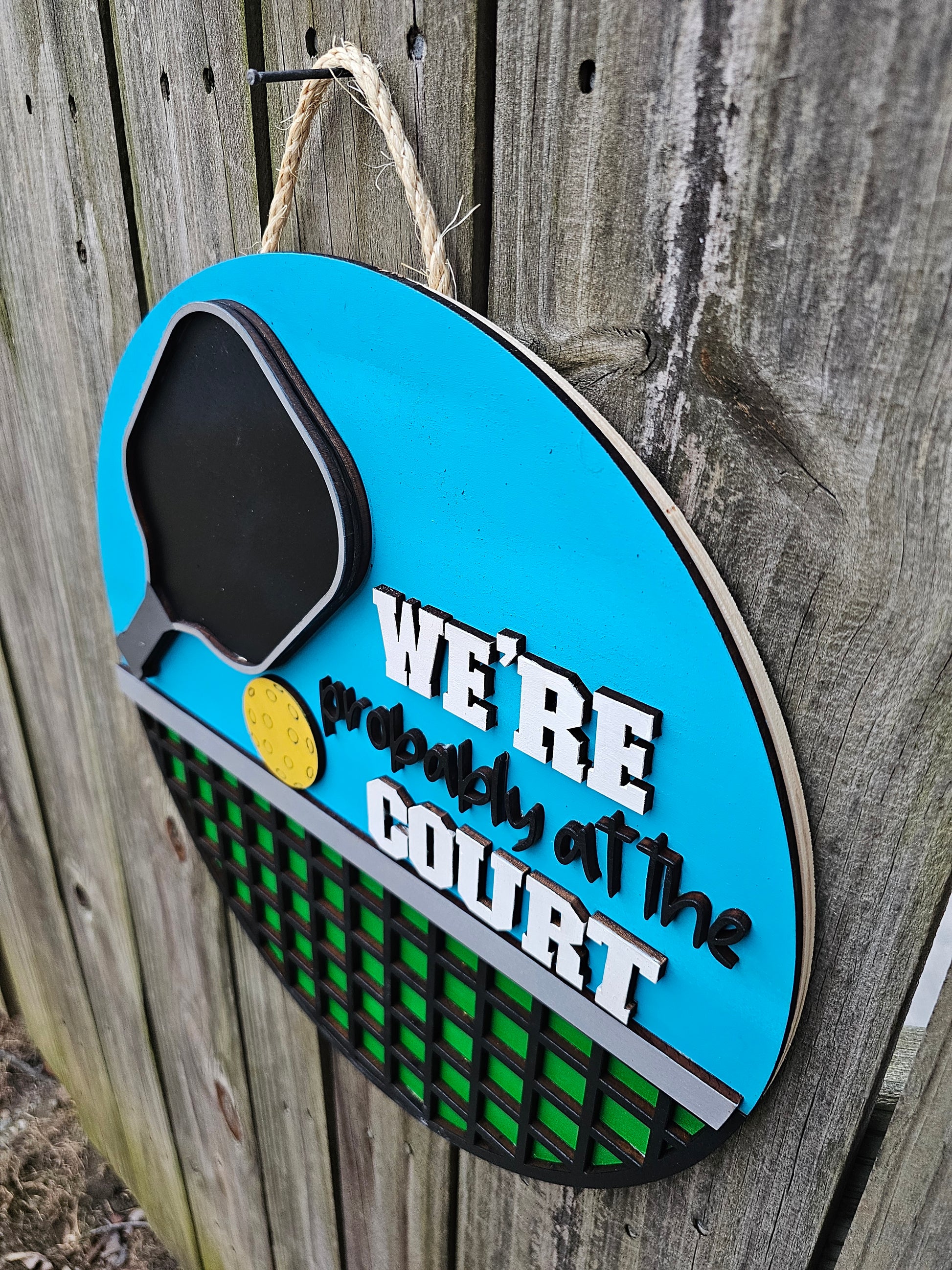Round wood pickleball themed door sign hanging on fence. The sign has a bright blue background on top with a pickleball paddle, ball, and "We're Probably At the Court." The bottom portion of the sign is green with black net. The sign is slightly angled in this photo showing 3D elements.