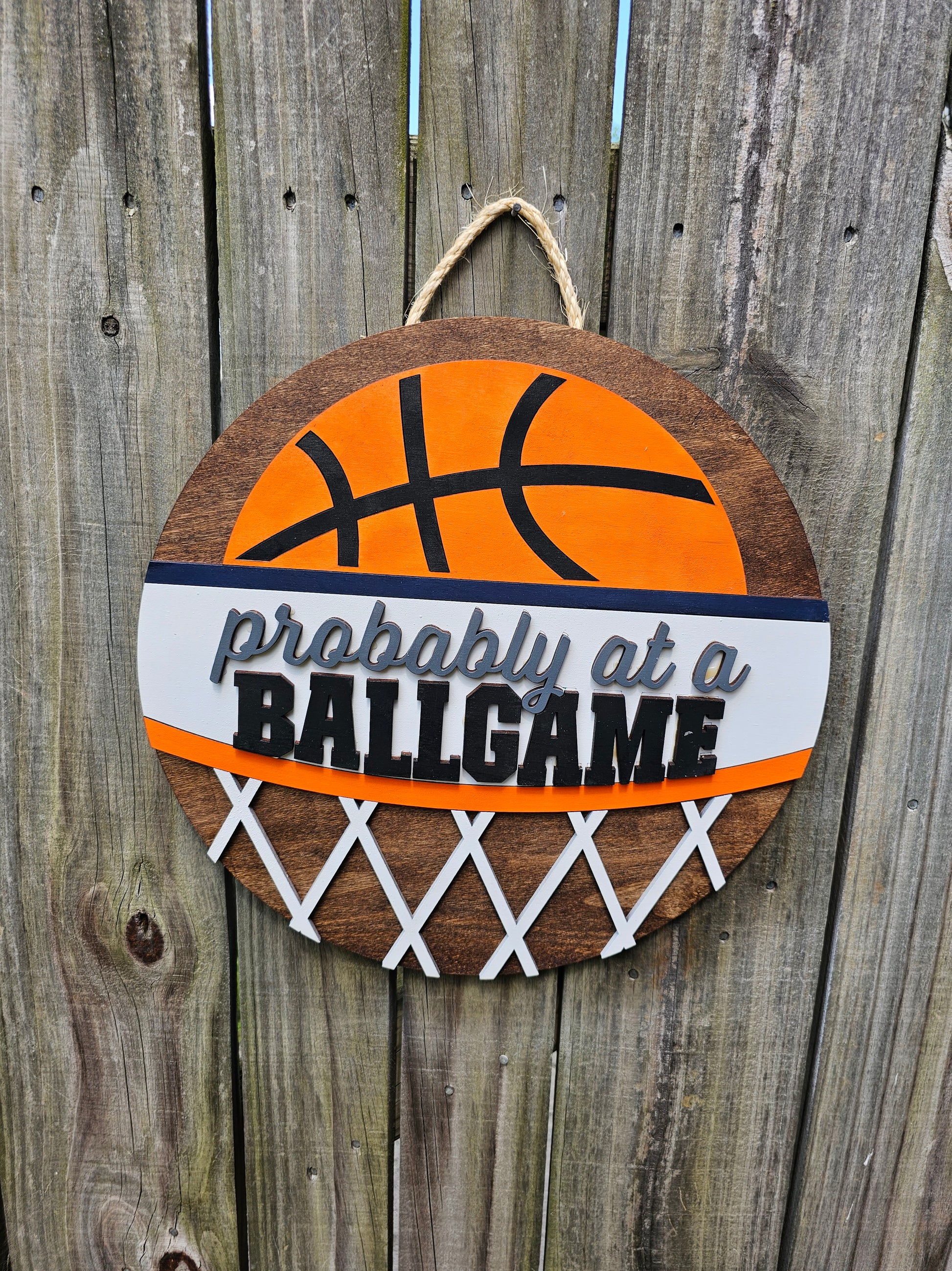 Round wood door sign with basketball design and text 'Probably at a BALLGAME'. Sign is hanging by twine on a wood fence. 