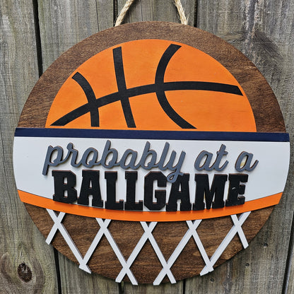 Round wood door sign with basketball design and text 'Probably at a BALLGAME'. Sign is hanging by twine on a wood fence. 