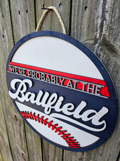 Round wood sign hanging on fence with "We're Probably At the Ballfield" and baseball laces. Sign is painted navy blue, red, and white. 