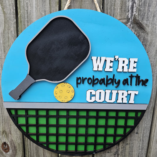 Round wood pickleball themed door sign hanging on fence. The sign has a bright blue background on top with a pickleball paddle, ball, and "We're Probably At the Court." The bottom portion of the sign is green with black net. The sign is front facing in this photo.