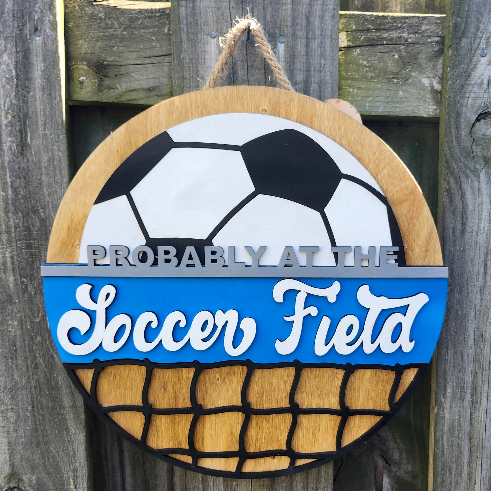 Round wood door sign made of wood. Stained background with black and white soccer ball at the top. Text "Probably at the Soccer Field" in gray and white. Blue background behind "soccer field". Black net at the bottom. Sign measures 14" in diameter and is hanging on a fence, front facing.