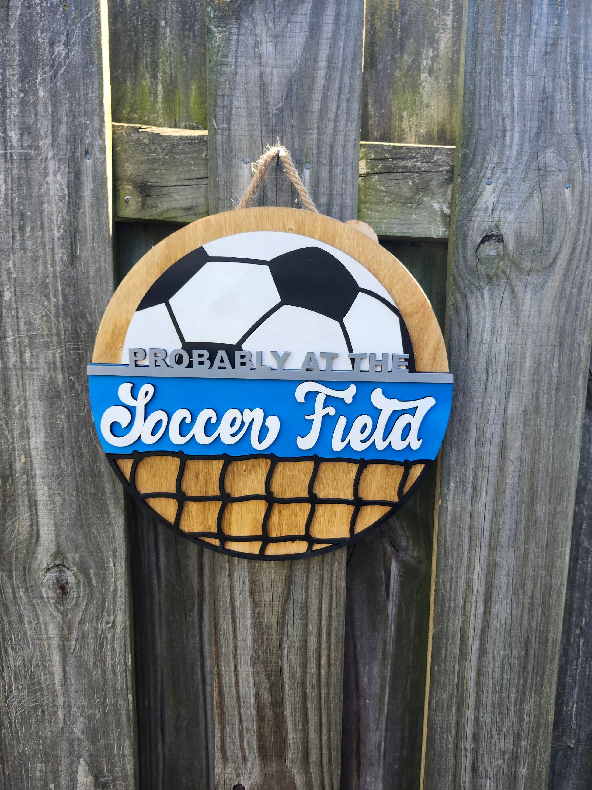 Round wood door sign made of wood. Stained background with black and white soccer ball at the top. Text "Probably at the Soccer Field" in gray and white. Blue background behind "soccer field". Black net at the bottom. Sign measures 14" in diameter and is hanging on a fence, front facing.