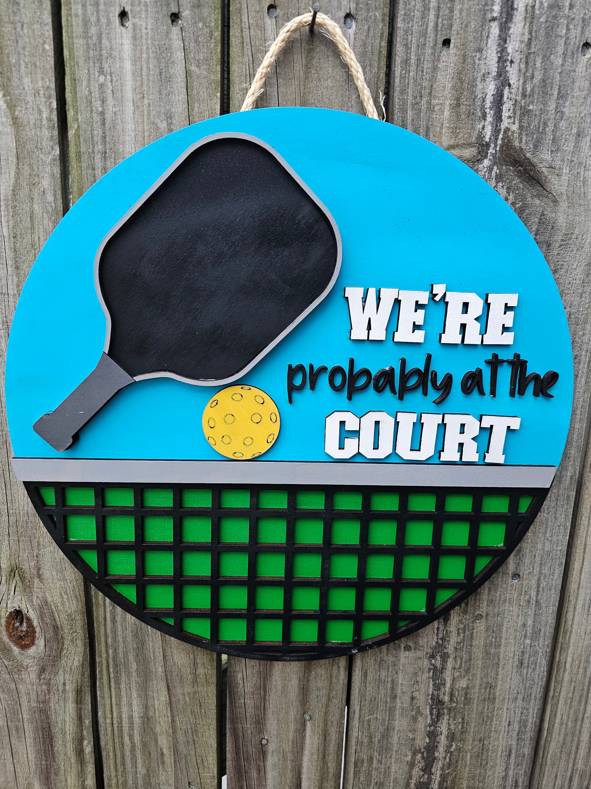 Round wood pickleball themed door sign hanging on fence. The sign has a bright blue background on top with a pickleball paddle, ball, and "We're Probably At the Court." The bottom portion of the sign is green with black net. The sign is facing forward in this photo.
