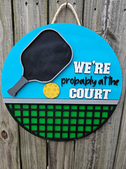 Round wood pickleball themed door sign hanging on fence. The sign has a bright blue background on top with a pickleball paddle, ball, and "We're Probably At the Court." The bottom portion of the sign is green with black net. The sign is facing forward in this photo.
