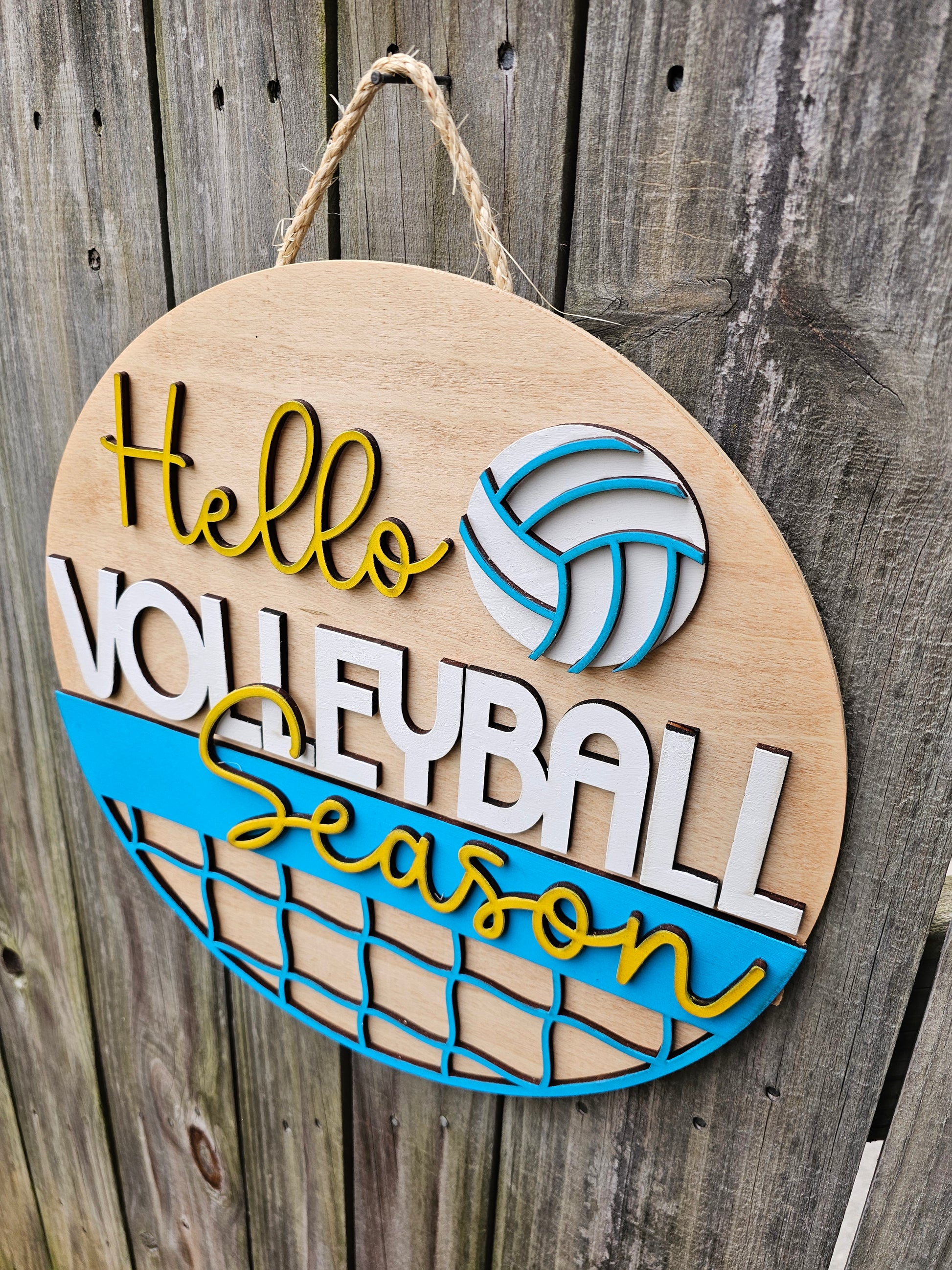Round wood door sign with light stained background and "Hello Volleyball Season" text. The sign also features a ball and net. The sign is hanging by twine on a fence. This photo is taken at an angle to show 3D elements on the sign.