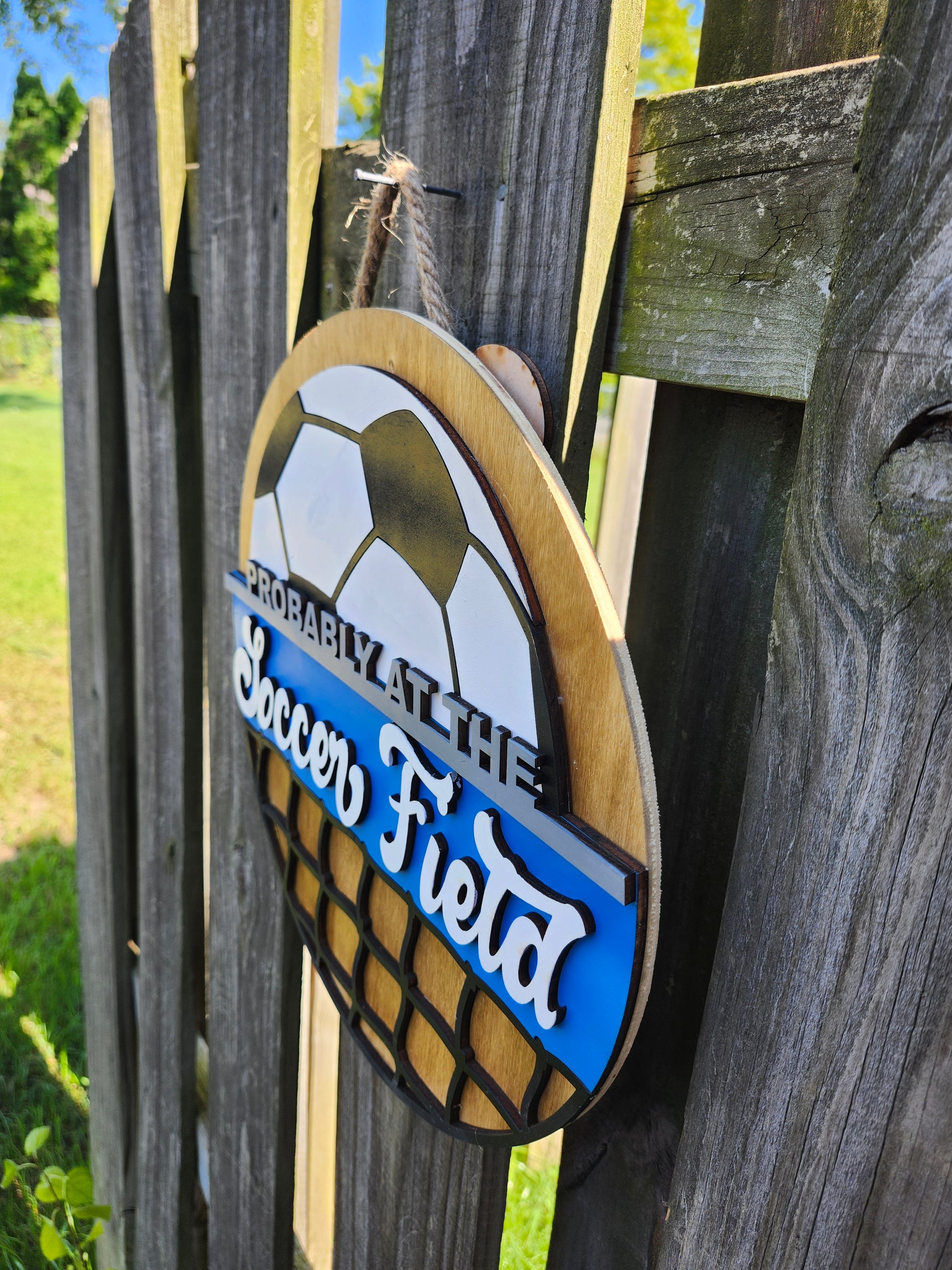 Round wood door sign made of wood. Stained background with black and white soccer ball at the top. Text "Probably at the Soccer Field" in gray and white. Blue background behind "soccer field". Black net at the bottom. Sign measures 14" in diameter and is hanging on a fence. This photo is taken on a slight angle showing that the sign has 3D dimensions. 