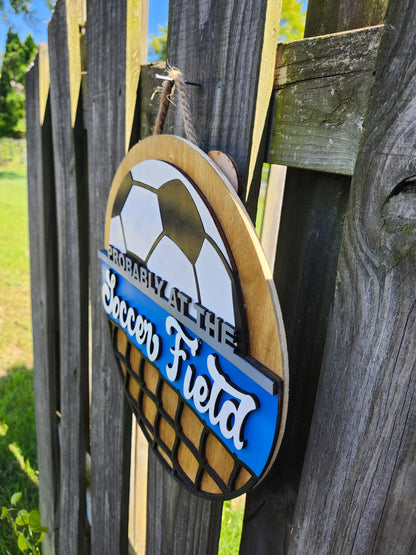 Round wood door sign made of wood. Stained background with black and white soccer ball at the top. Text "Probably at the Soccer Field" in gray and white. Blue background behind "soccer field". Black net at the bottom. Sign measures 14" in diameter and is hanging on a fence. This photo is taken on a slight angle showing that the sign has 3D dimensions. 