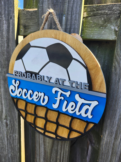Round wood door sign made of wood. Stained background with black and white soccer ball at the top. Text "Probably at the Soccer Field" in gray and white. Blue background behind "soccer field". Black net at the bottom. Sign measures 14" in diameter and is hanging on a fence. This photo is taken on a slight angle showing that the sign has 3D dimensions. 