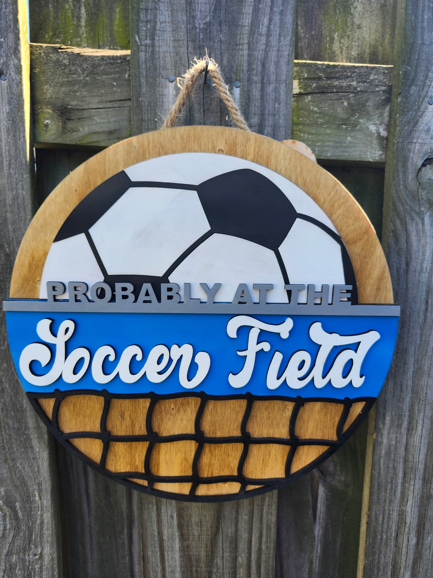 Round wood door sign made of wood. Stained background with black and white soccer ball at the top. Text "Probably at the Soccer Field" in gray and white. Blue background behind "soccer field". Black net at the bottom. Sign measures 14" in diameter and is hanging on a fence, front facing.
