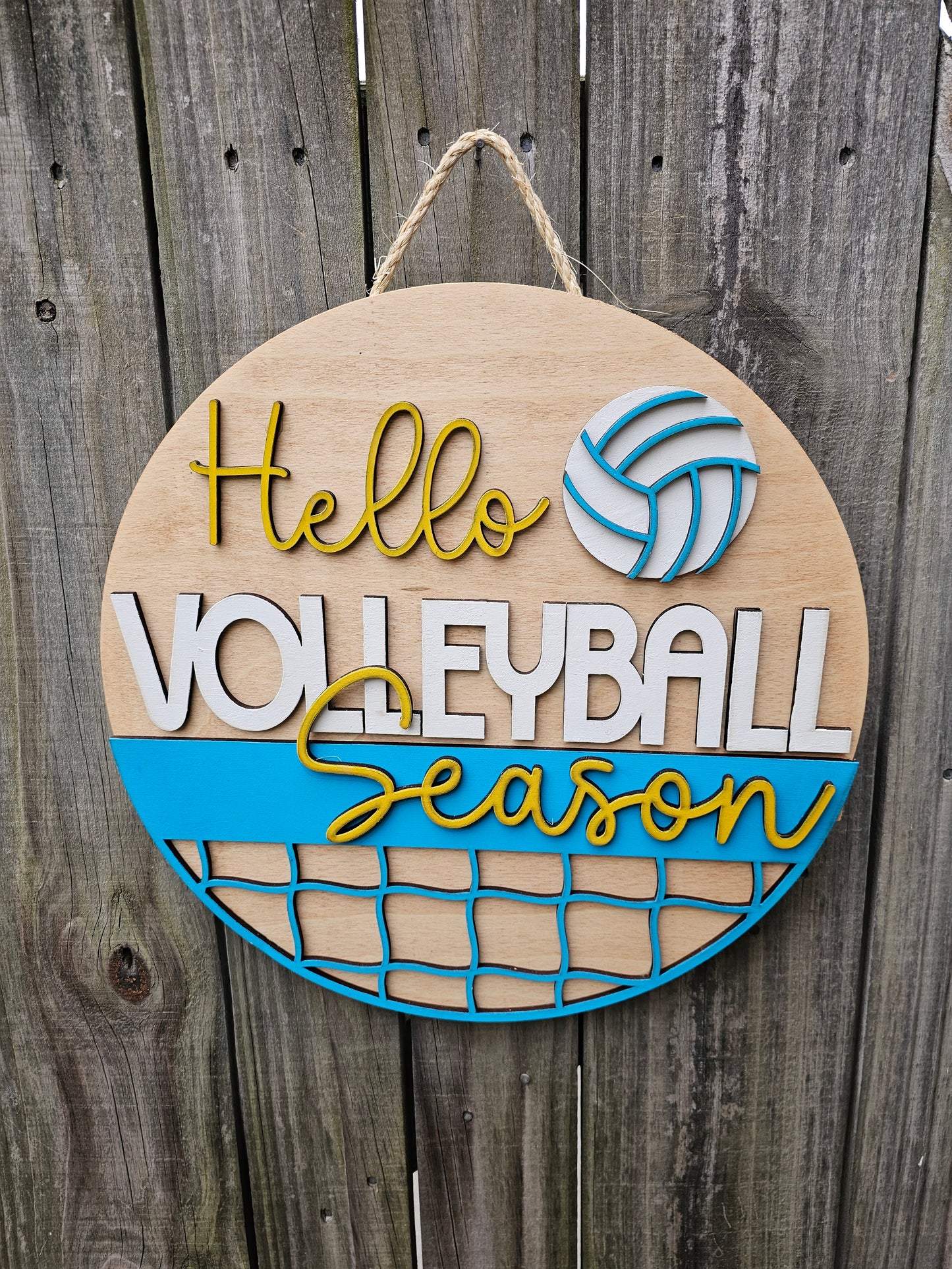 Round wood door sign with light stained background and "Hello Volleyball Season" text. The sign also features a ball and net. The sign is hanging by twine on a fence.