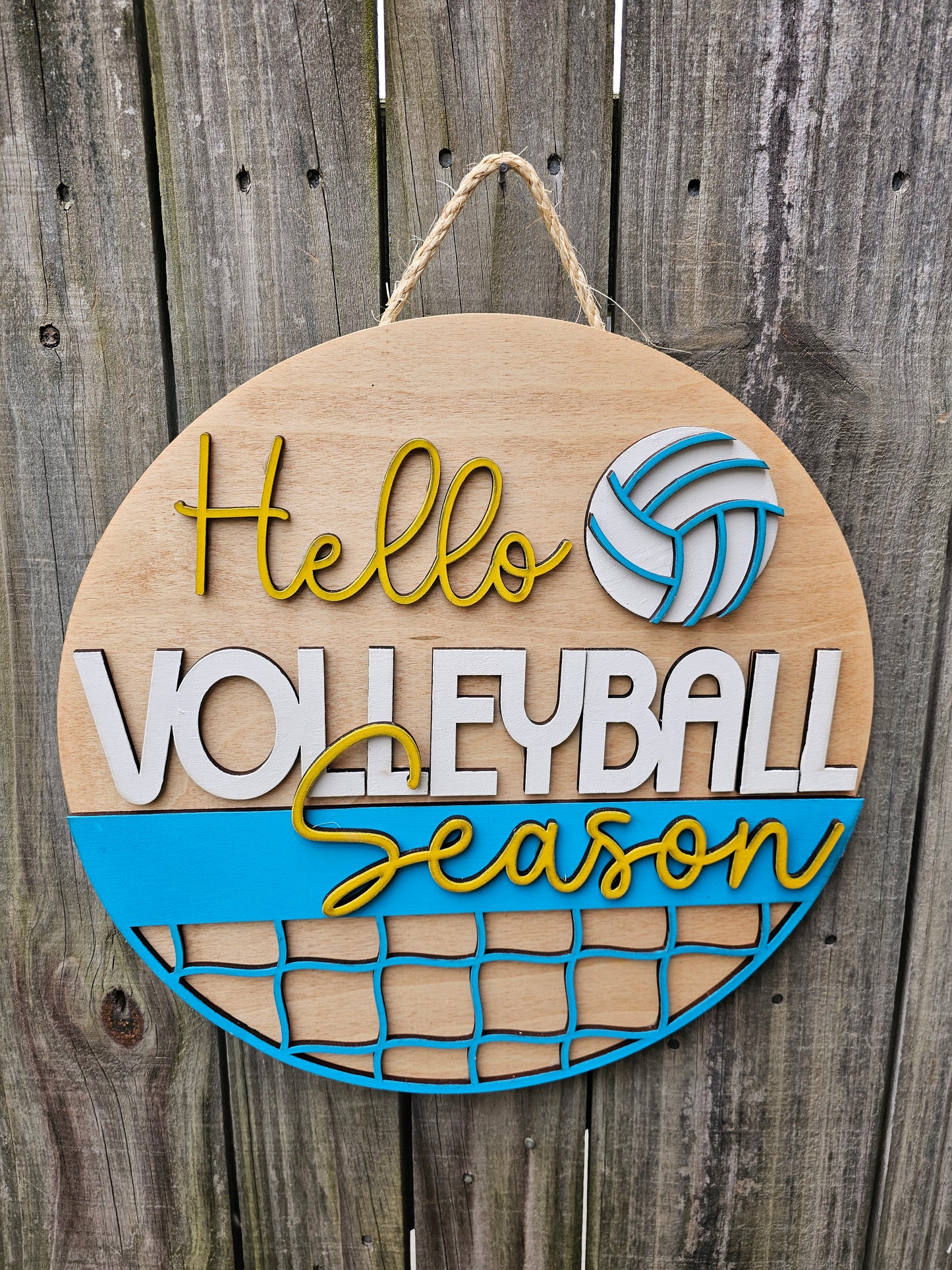 Round wood door sign with light stained background and "Hello Volleyball Season" text. The sign also features a ball and net. The sign is hanging by twine on a fence.