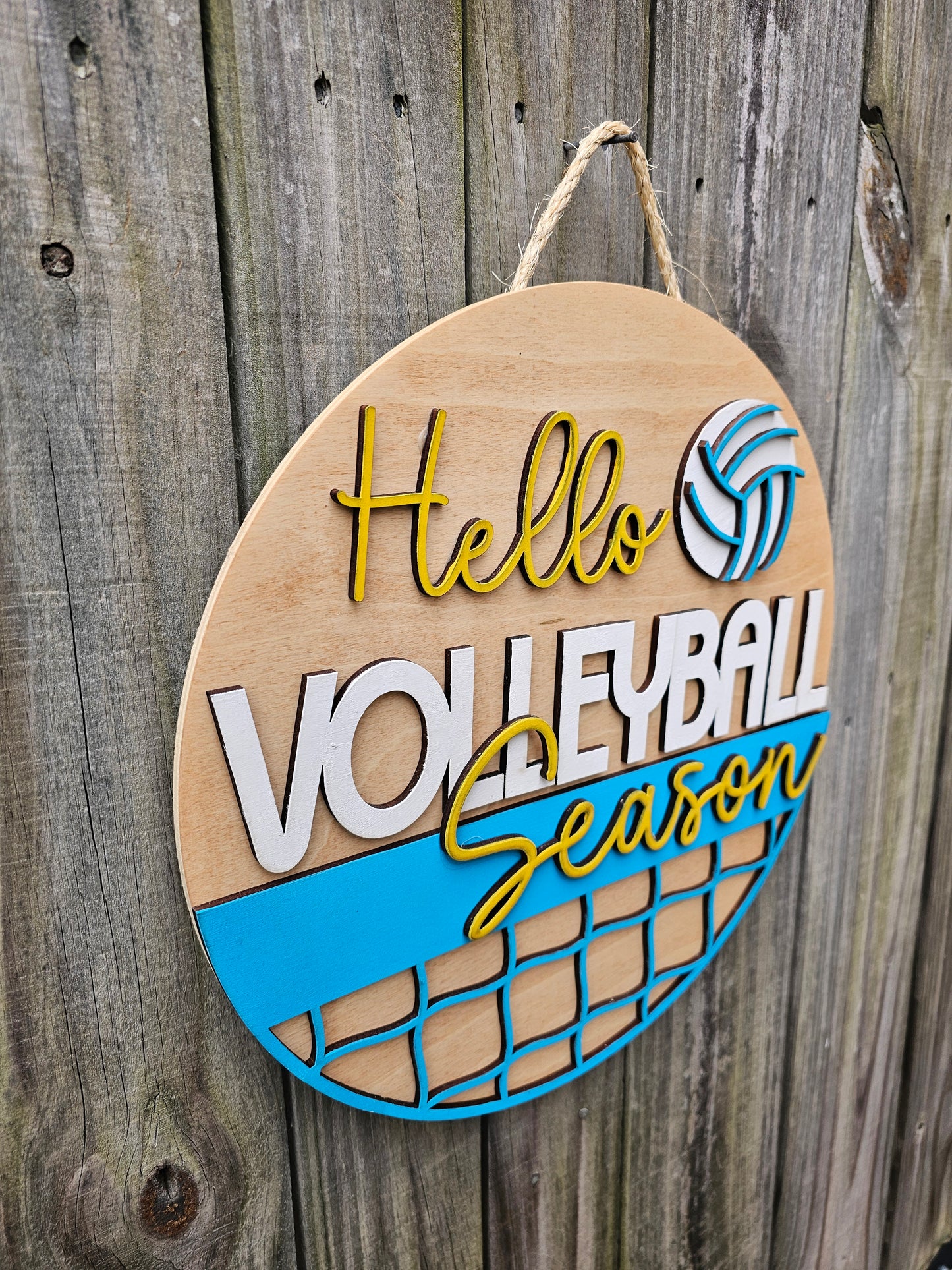 Round wood door sign with light stained background and "Hello Volleyball Season" text. The sign also features a ball and net. The sign is hanging by twine on a fence. This photo is taken at an angle to show 3D elements on the sign.