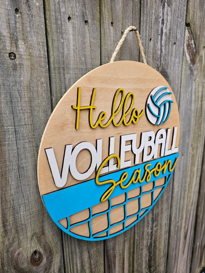 Round wood door sign with light stained background and "Hello Volleyball Season" text. The sign also features a ball and net. The sign is hanging by twine on a fence. This photo is taken at an angle to show 3D elements on the sign.