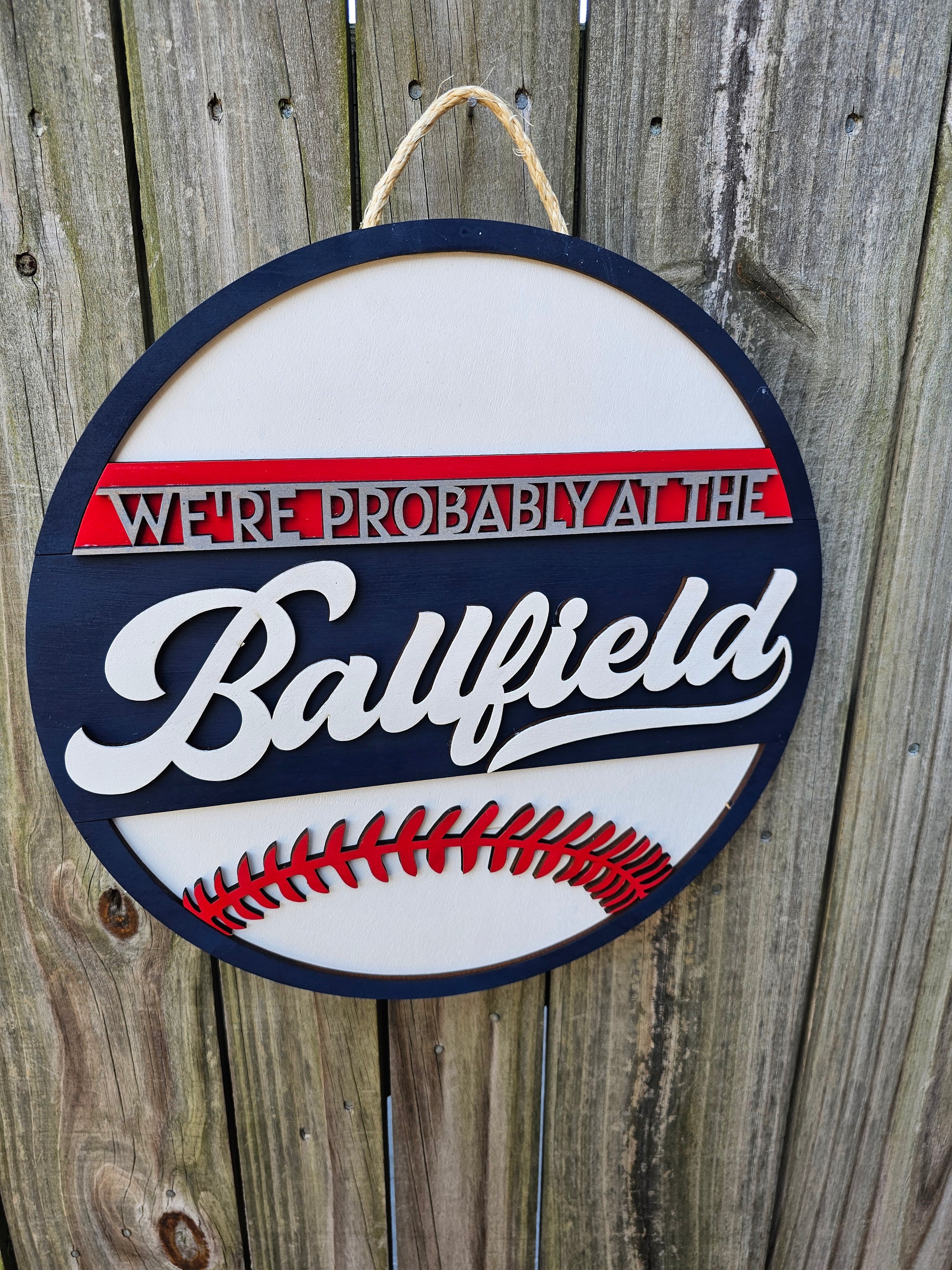 Round wood sign hanging on fence with "We're Probably At the Ballfield" and baseball laces. Sign is painted navy blue, red, and white.