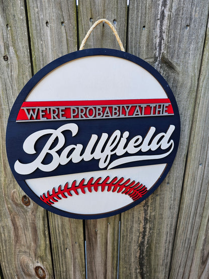 Round wood sign hanging on fence with "We're Probably At the Ballfield" and baseball laces. Sign is painted navy blue, red, and white.