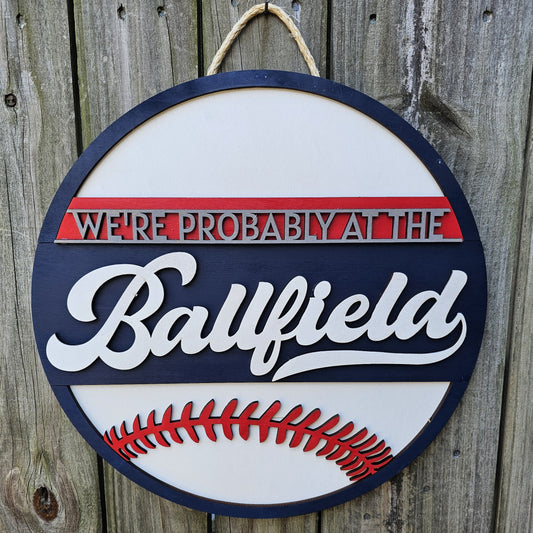 Round wood sign hanging on fence with "We're Probably At the Ballfield" and baseball laces. Sign is painted navy blue, red, and white. In this photo the sign is forward facing.