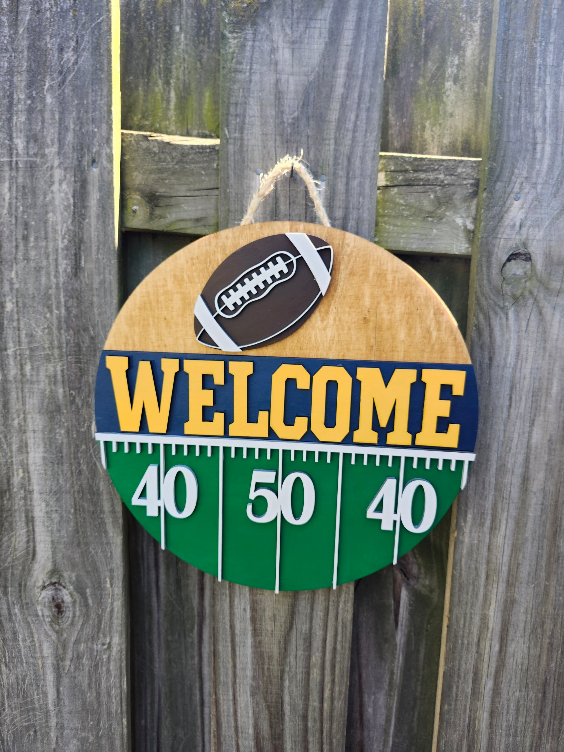 Round wood door sign hanging from a fence with twine. The sign's background has a light colored stain. At the top of the sign is a brown football with white laces. Below that is "Welcome" in bold yellow letters with a navy blue background. Below that (at the bottom of the sign) is a green semi-circle with white yard lines marking "40", "50", "40". This sign is facing directly forward in this photo.