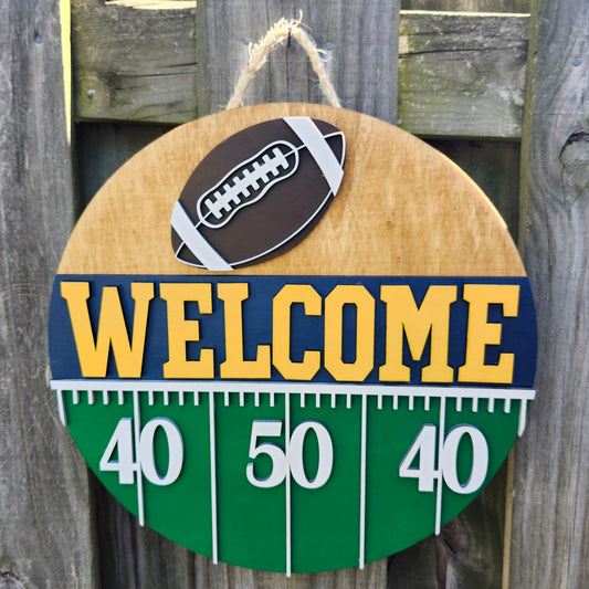 Round wood door sign hanging from a fence with twine. The sign's background has a light colored stain. At the top of the sign is a brown football with white laces. Below that is "Welcome" in bold yellow letters with a navy blue background. Below that (at the bottom of the sign) is a green semi-circle with white yard lines marking "40", "50", "40". This sign is facing directly forward in this photo.