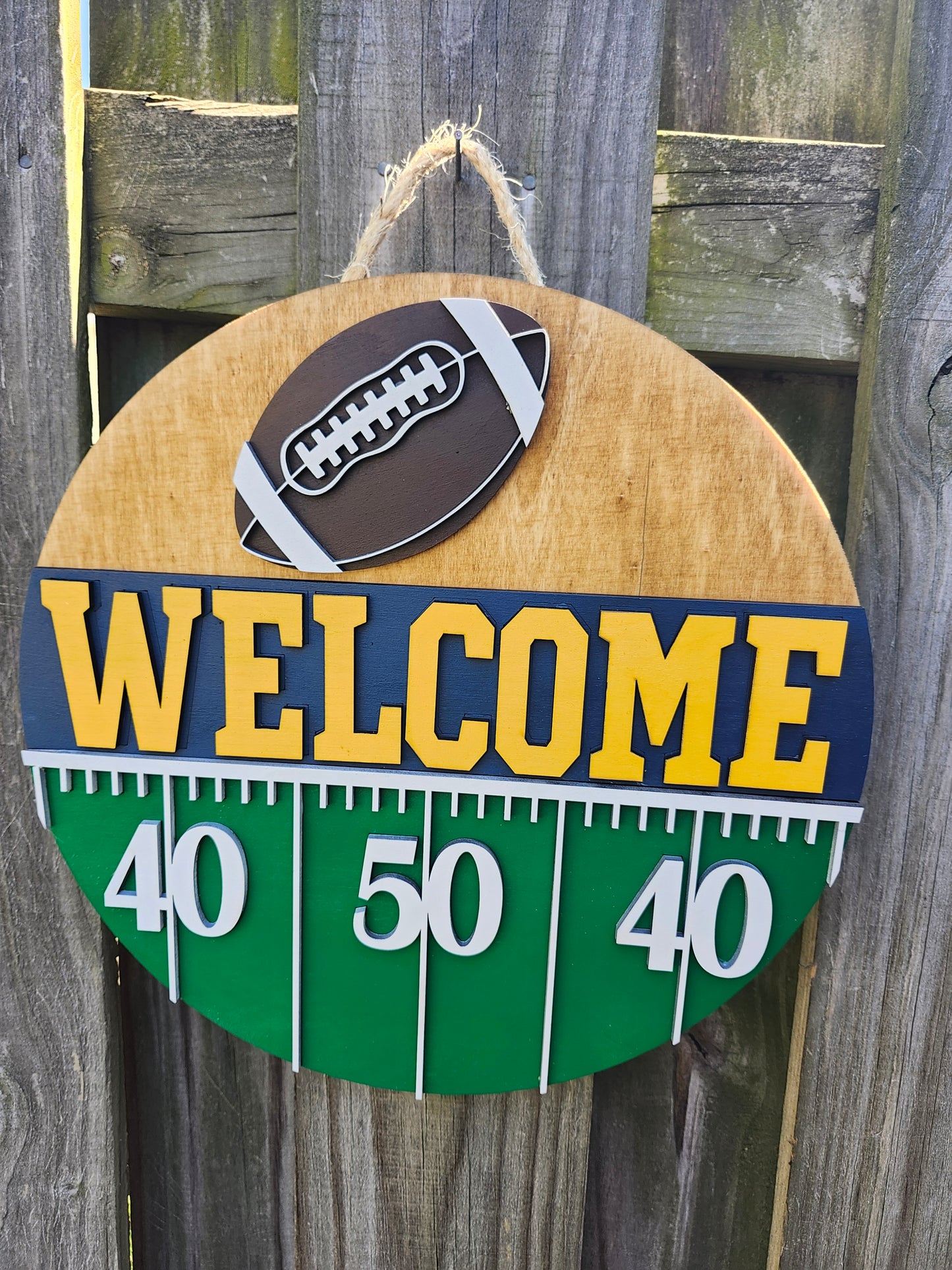 Round wood door sign hanging from a fence with twine. The sign's background has a light colored stain. At the top of the sign is a brown football with white laces. Below that is "Welcome" in bold yellow letters with a navy blue background. Below that (at the bottom of the sign) is a green semi-circle with white yard lines marking "40", "50", "40". This sign is from a slightly off-center angle.