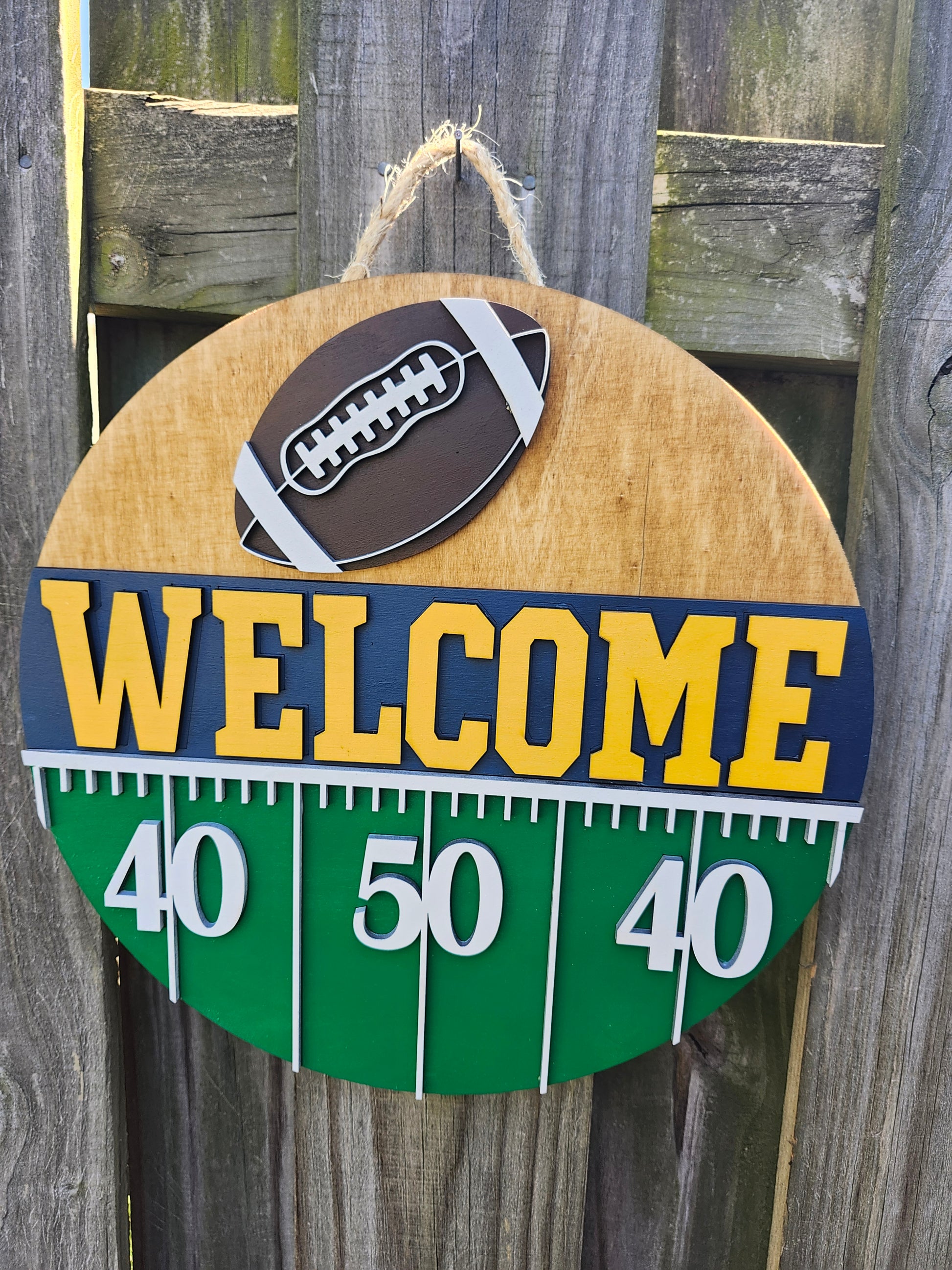 Round wood door sign hanging from a fence with twine. The sign's background has a light colored stain. At the top of the sign is a brown football with white laces. Below that is "Welcome" in bold yellow letters with a navy blue background. Below that (at the bottom of the sign) is a green semi-circle with white yard lines marking "40", "50", "40". This sign is from a slightly off-center angle.