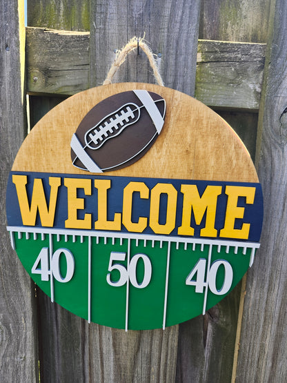 Round wood door sign hanging from a fence with twine. The sign's background has a light colored stain. At the top of the sign is a brown football with white laces. Below that is "Welcome" in bold yellow letters with a navy blue background. Below that (at the bottom of the sign) is a green semi-circle with white yard lines marking "40", "50", "40". This sign is from a slightly off-center angle.