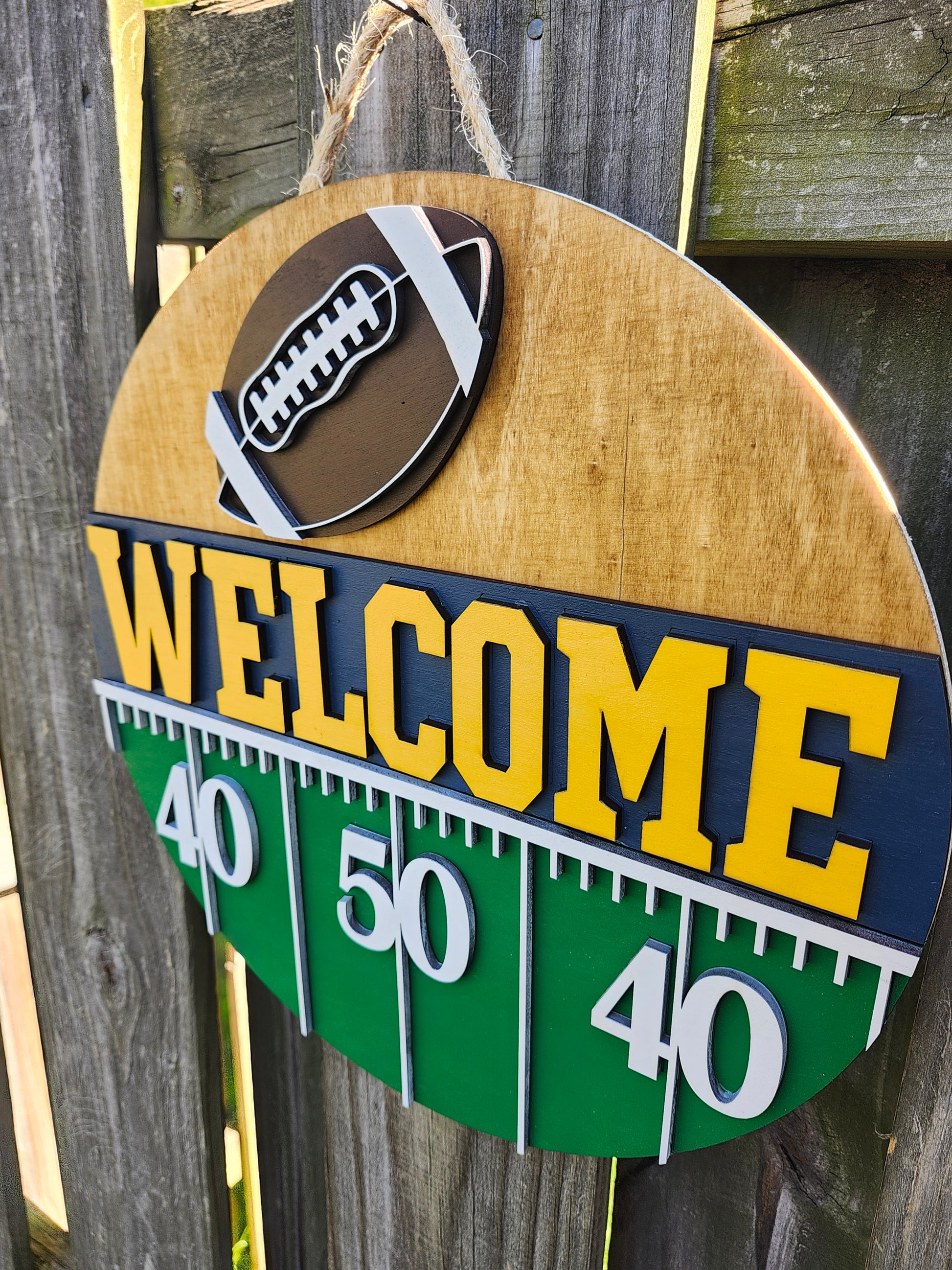 Round wood door sign hanging from a fence with twine. The sign's background has a light colored stain. At the top of the sign is a brown football with white laces. Below that is "Welcome" in bold yellow letters with a navy blue background. Below that (at the bottom of the sign) is a green semi-circle with white yard lines marking "40", "50", "40". The sign is at a slight angle at this photo showing the 3D dimension.