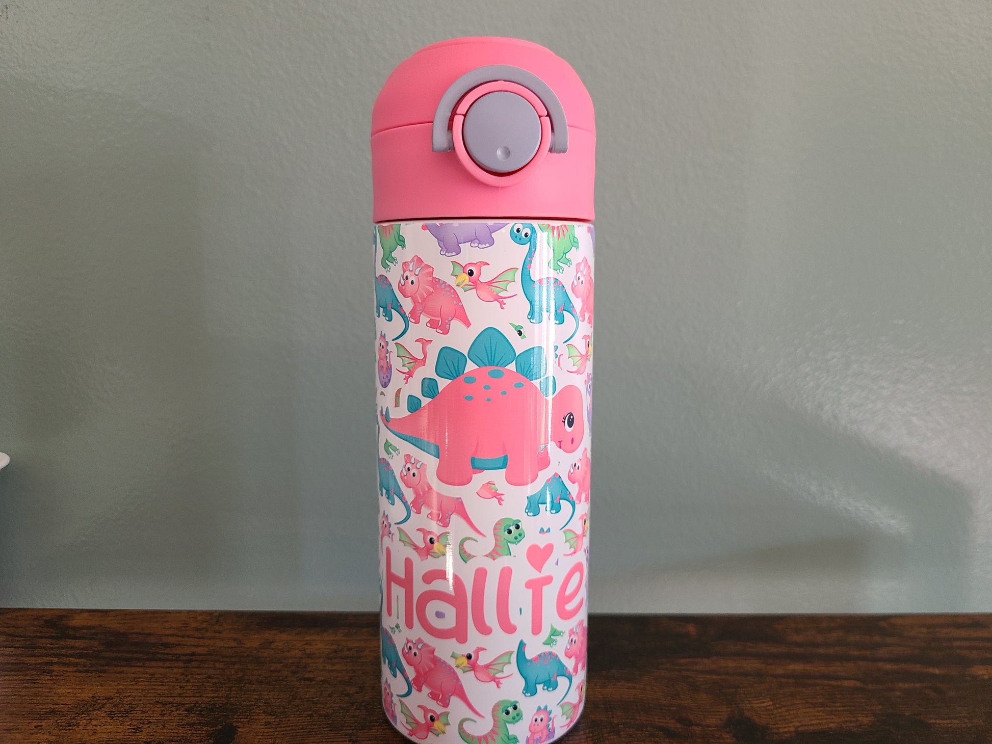 12 oz water bottle with pink lid that has a built in straw, locking push button and handle. This design features pastel rainbow dinosaurs. Personalized in coordinating colors. Back to School.