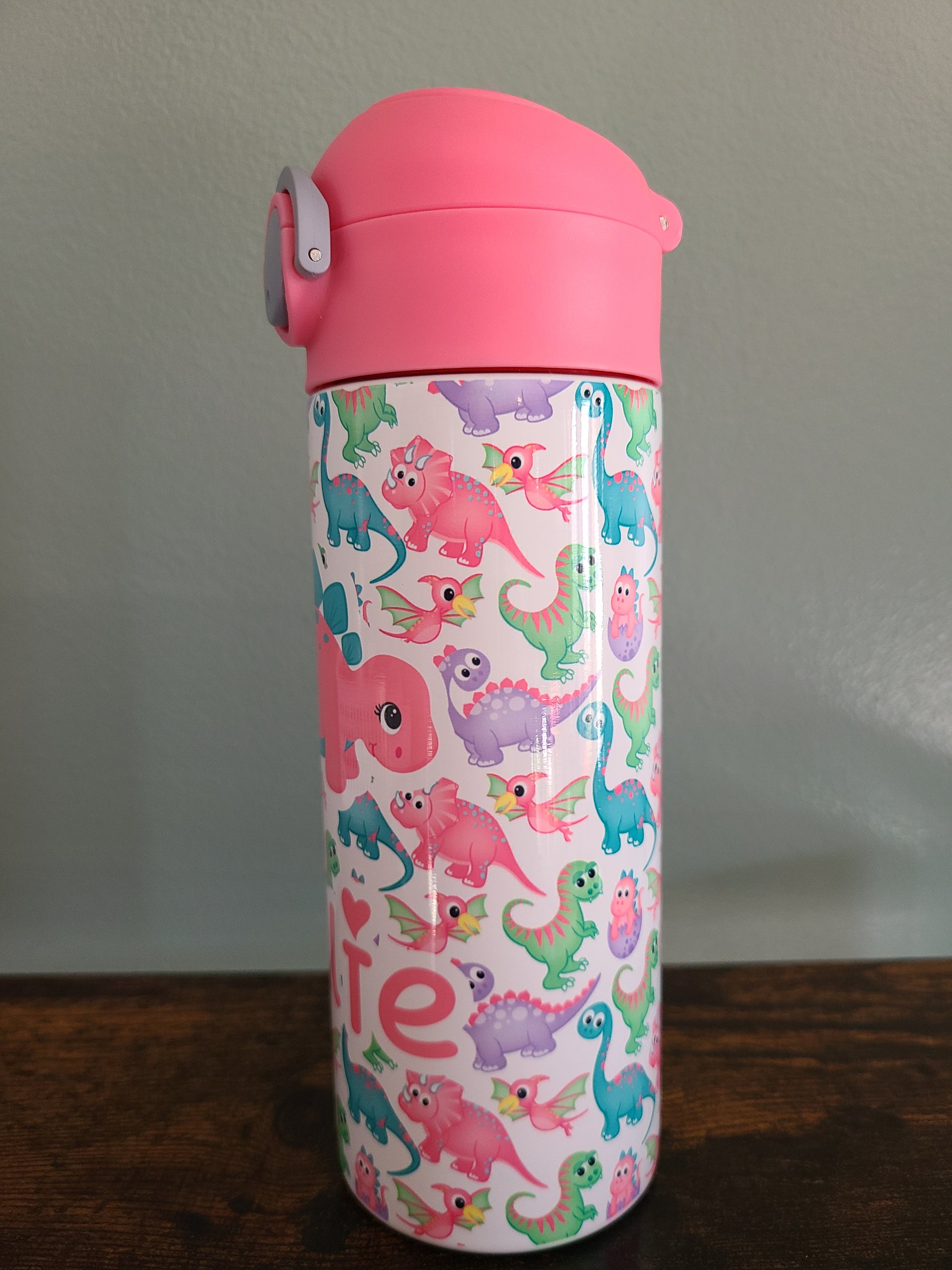 12 oz water bottle with pink lid that has a built in straw, locking push button and handle. This design features pastel rainbow dinosaurs. Personalized in coordinating colors. Back to School.