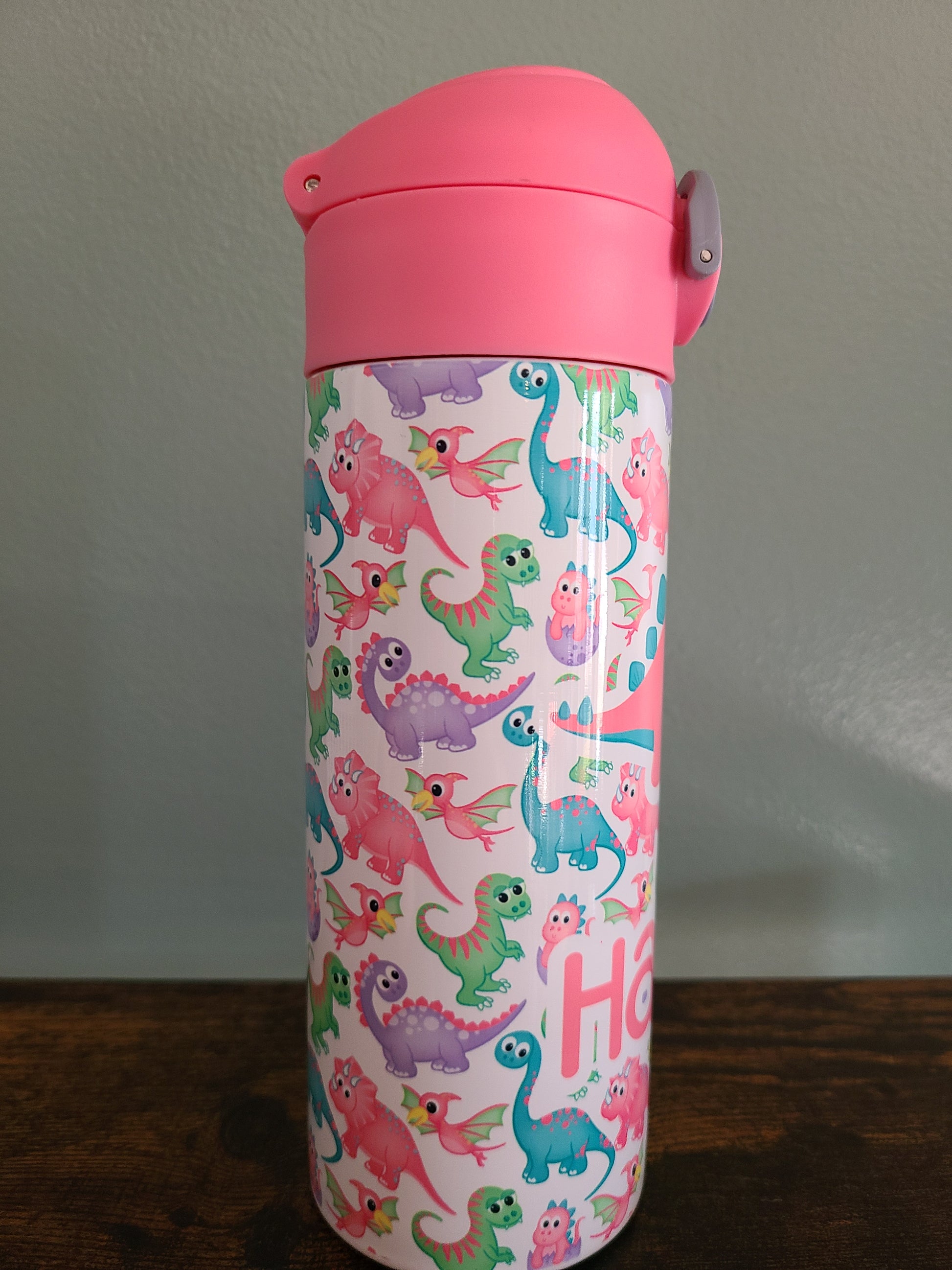 12 oz water bottle with pink lid that has a built in straw, locking push button and handle. This design features pastel rainbow dinosaurs. Personalized in coordinating colors. Back to School.