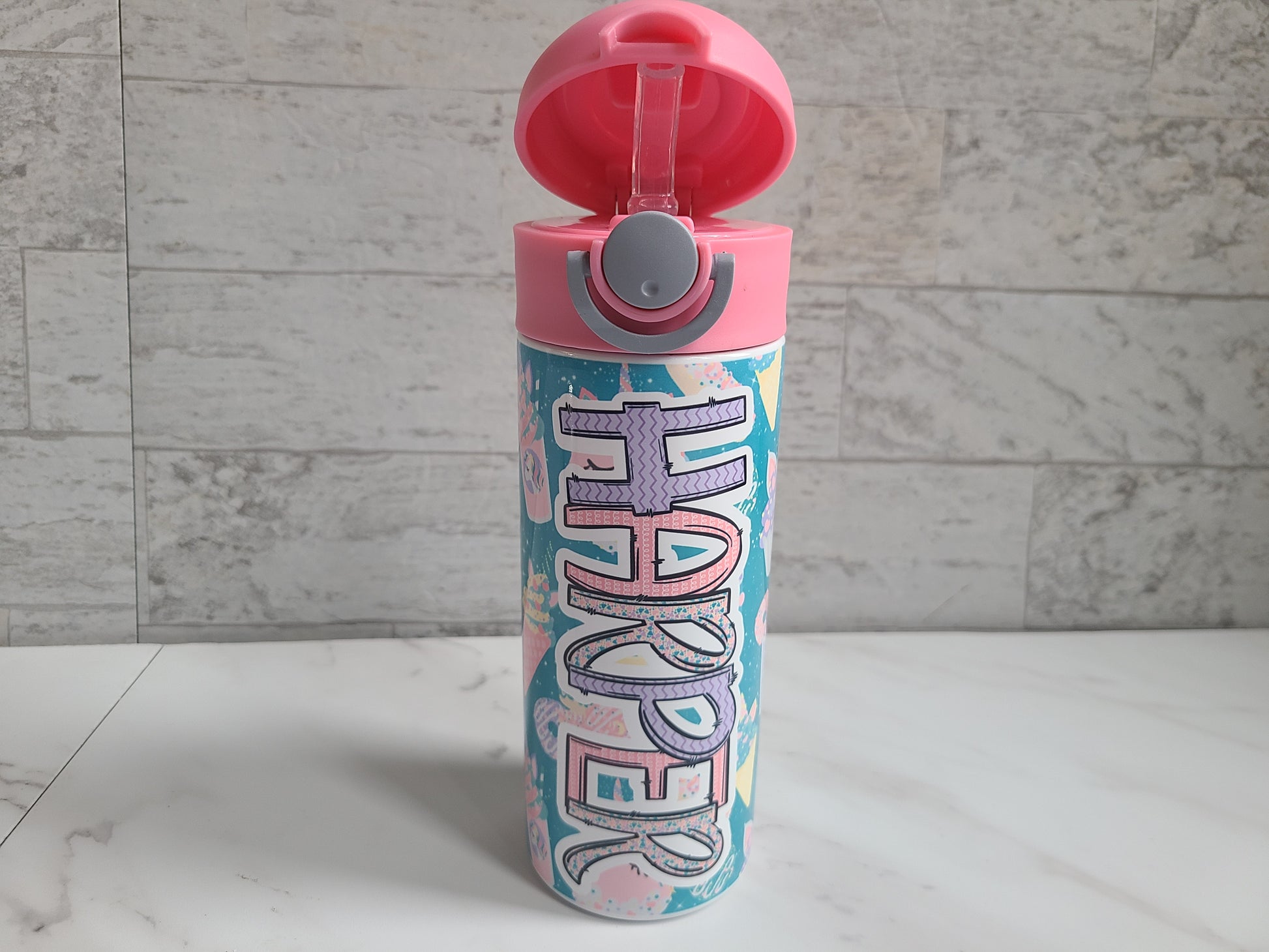 12 oz water bottle with pink lid that has a built-in straw, locking push button and handle. This design features pastel unicorn cupcakes and ice cream cones. Personalized in coordinating colors. Lid is popped open easily to show the silicone straw