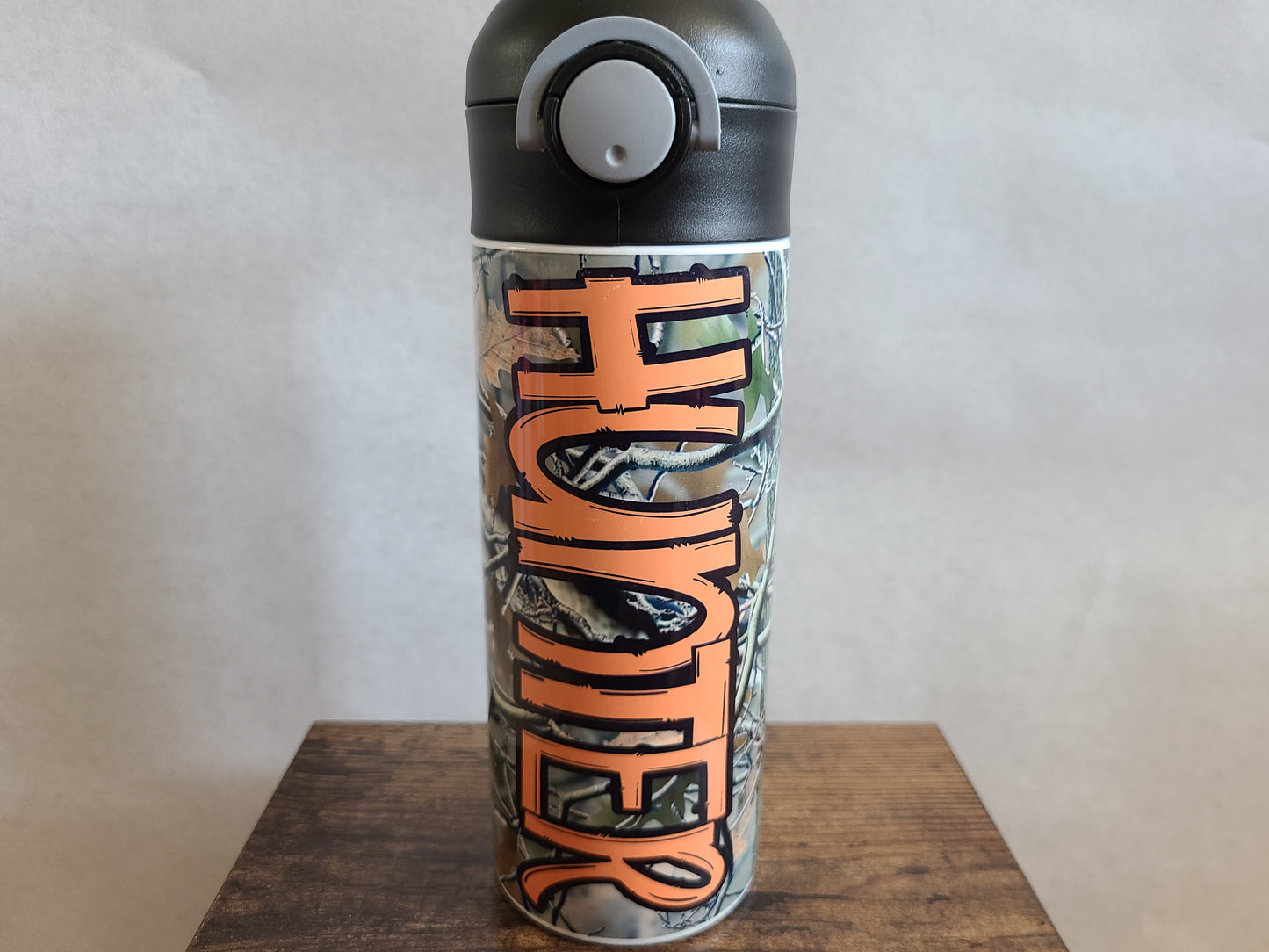 12 oz water bottle with a black lid that has a built-in straw, locking push button and handle. This design features real tree camo with orange personalization. Back to School.